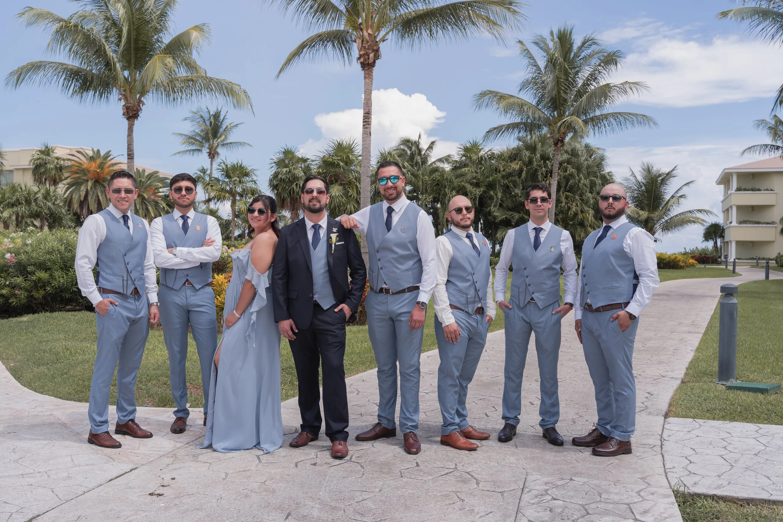 A group of nine people dressed in formal attire, standing outdoors near palm trees on a sunny day, in what appears to be a wedding or special event setting.