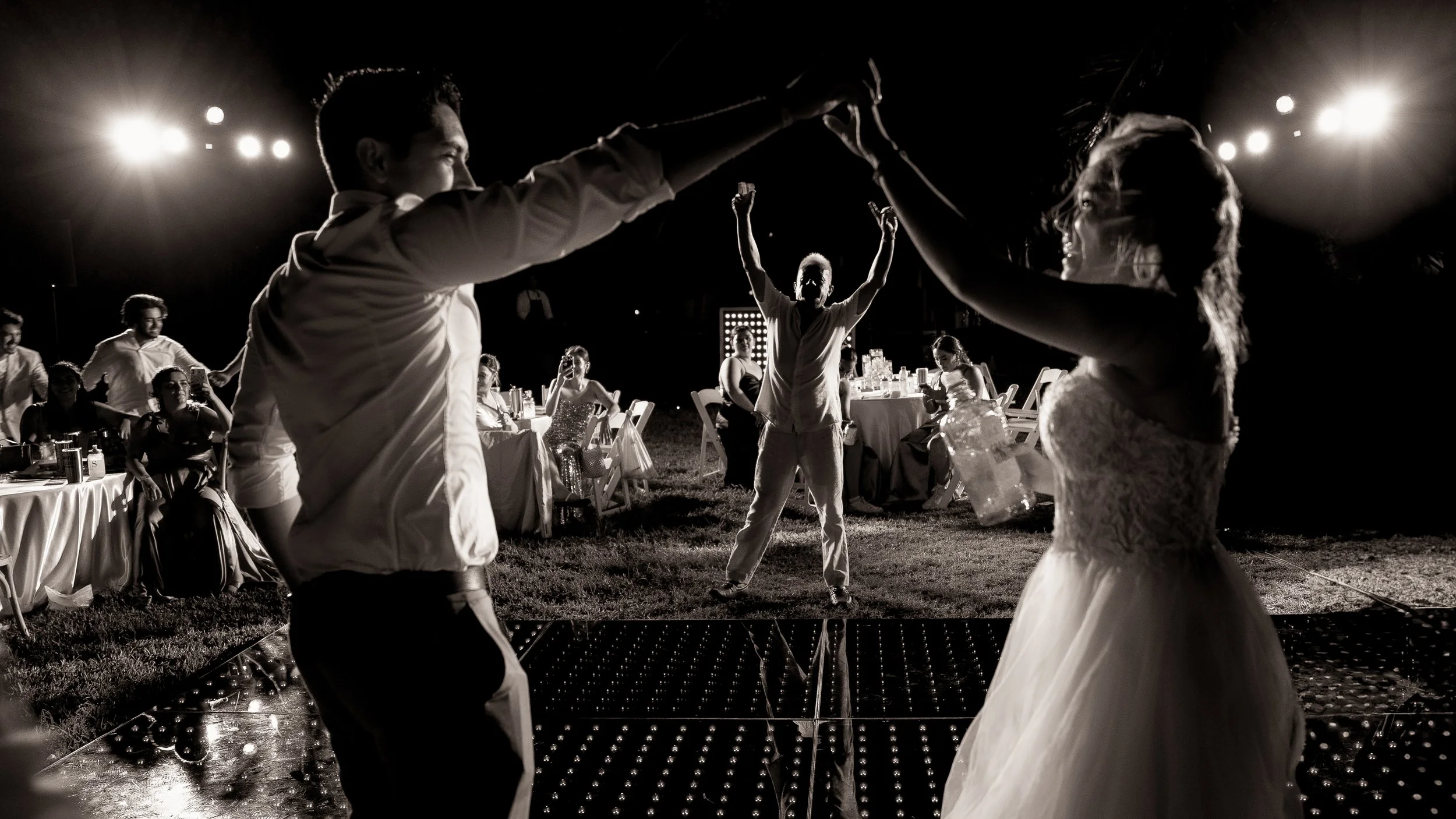 A black and white photo of a dance floor at night, with a man and woman dancing in the foreground, the couple holding hands. In the background, a person with arms raised, others sitting at tables, and bright lights overhead illuminating the scene.