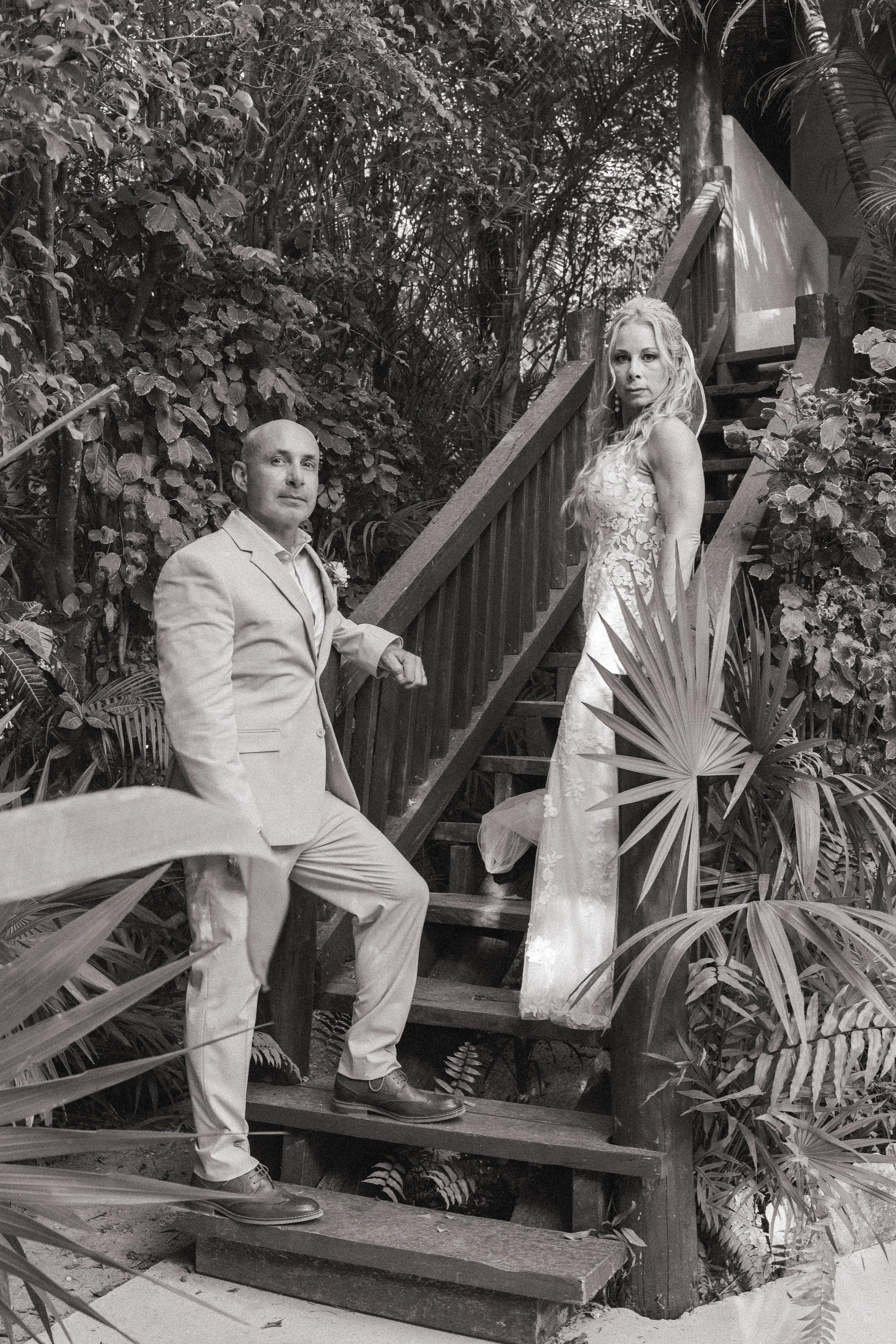 A black and white photo of a man in a suit and a woman in a wedding dress on a wooden staircase surrounded by tropical plants.