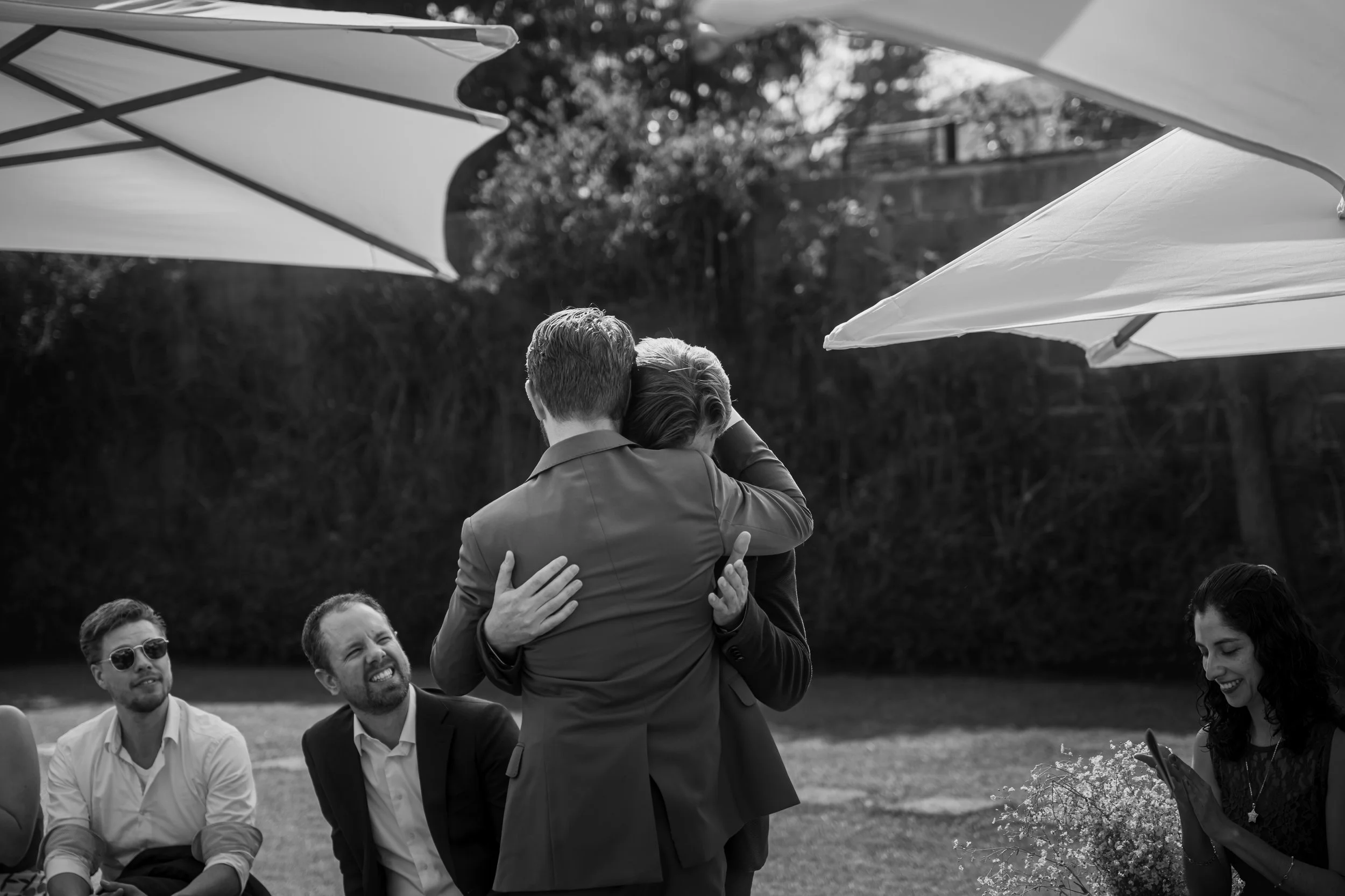 Two men embrace in a hug at an outdoor gathering, with other people sitting nearby and smiling, under large umbrellas in a garden setting.