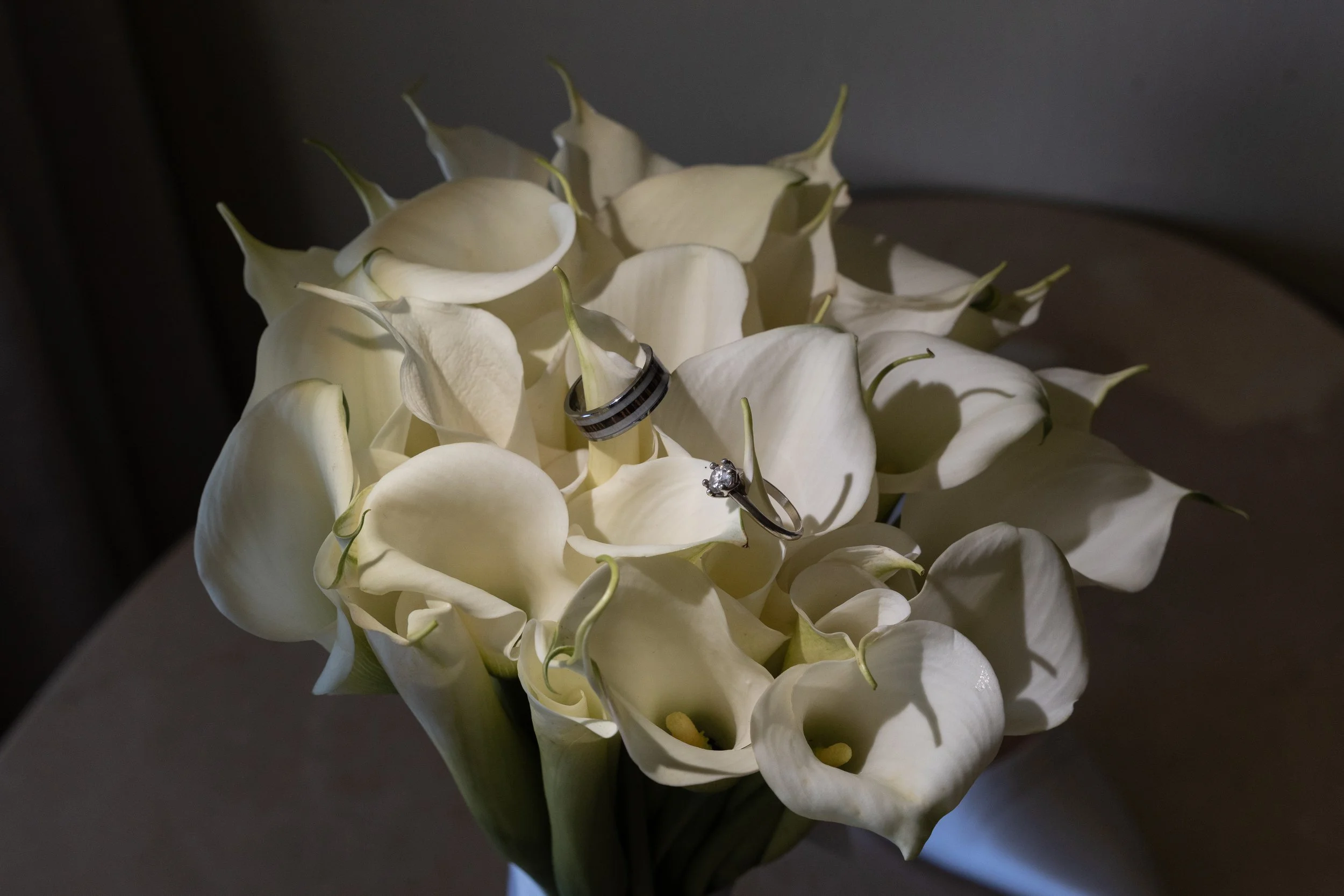 A bouquet of white calla lilies with a set of wedding rings placed on top, one ring with a diamond stone.