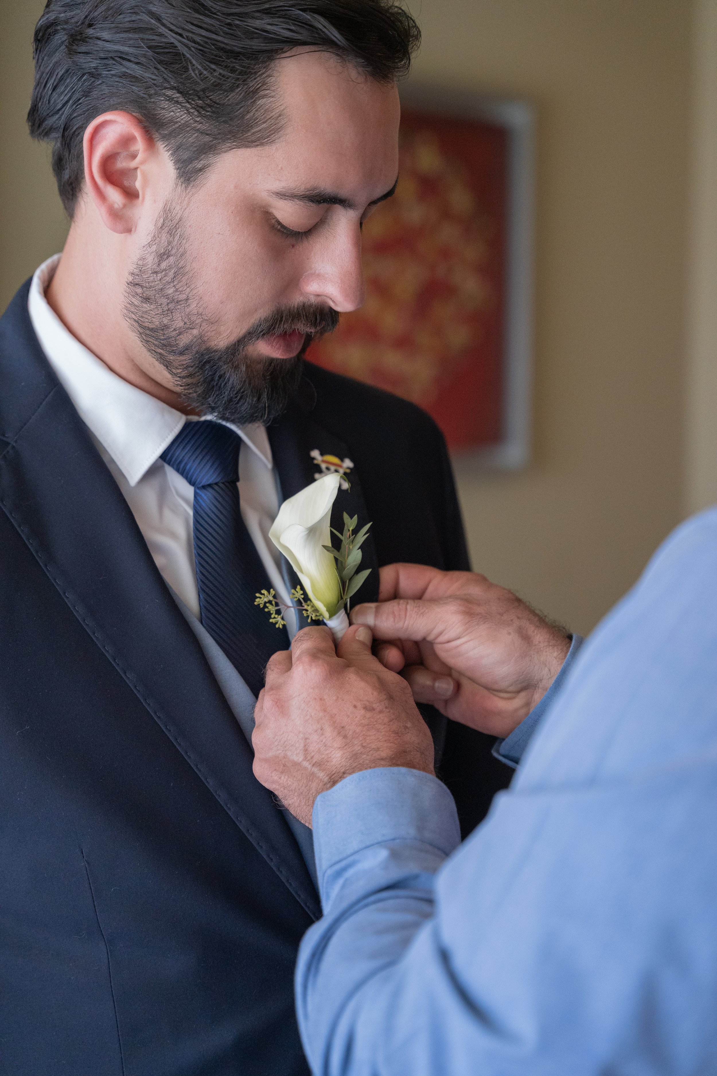 A man in a suit is having a boutonniere pinning ceremony with another man. The man in the suit has dark hair and a beard.