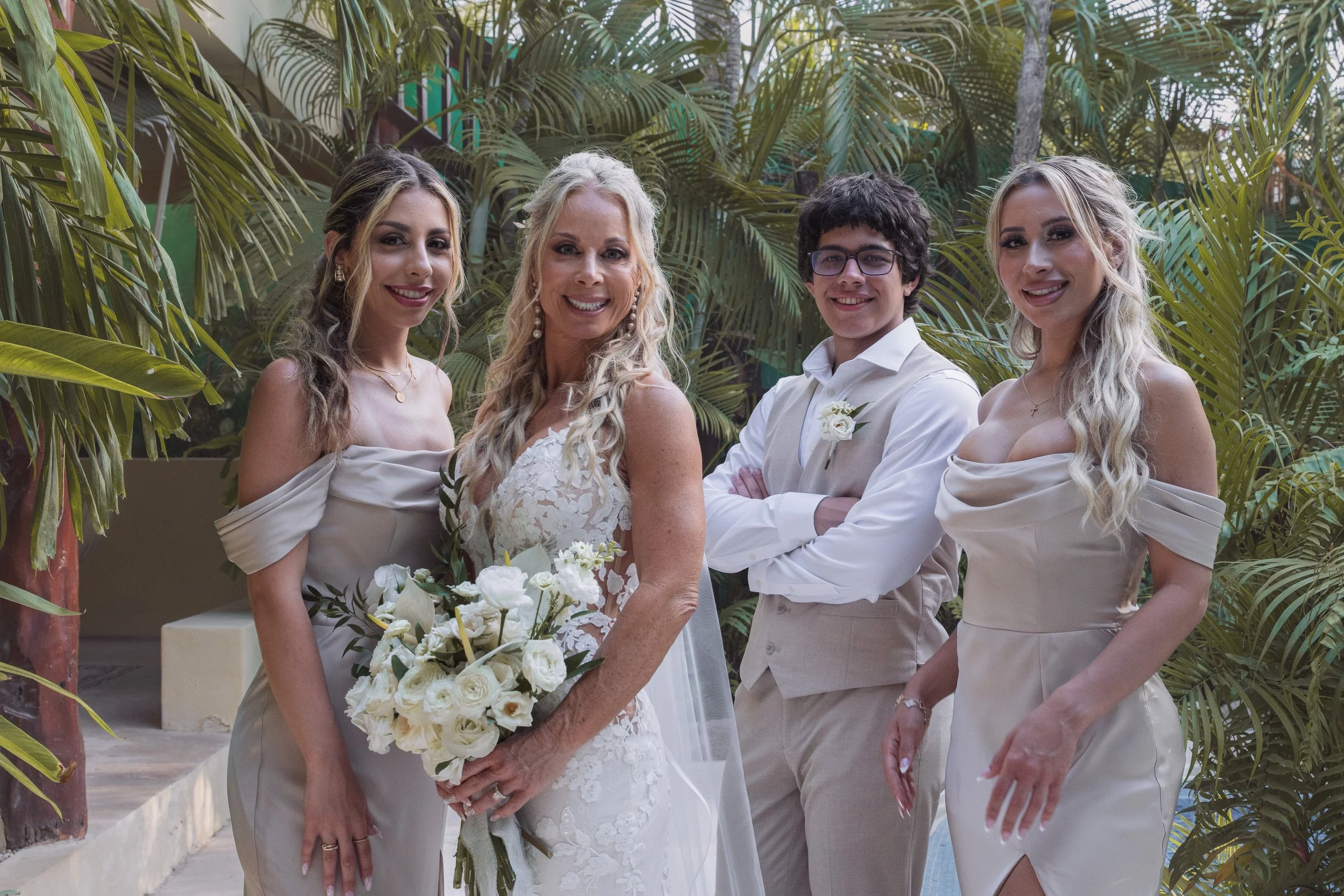 A woman in a white lace wedding dress holding a bouquet of white flowers is surrounded by three women and one man, all dressed in formal attire, in a lush, tropical setting.