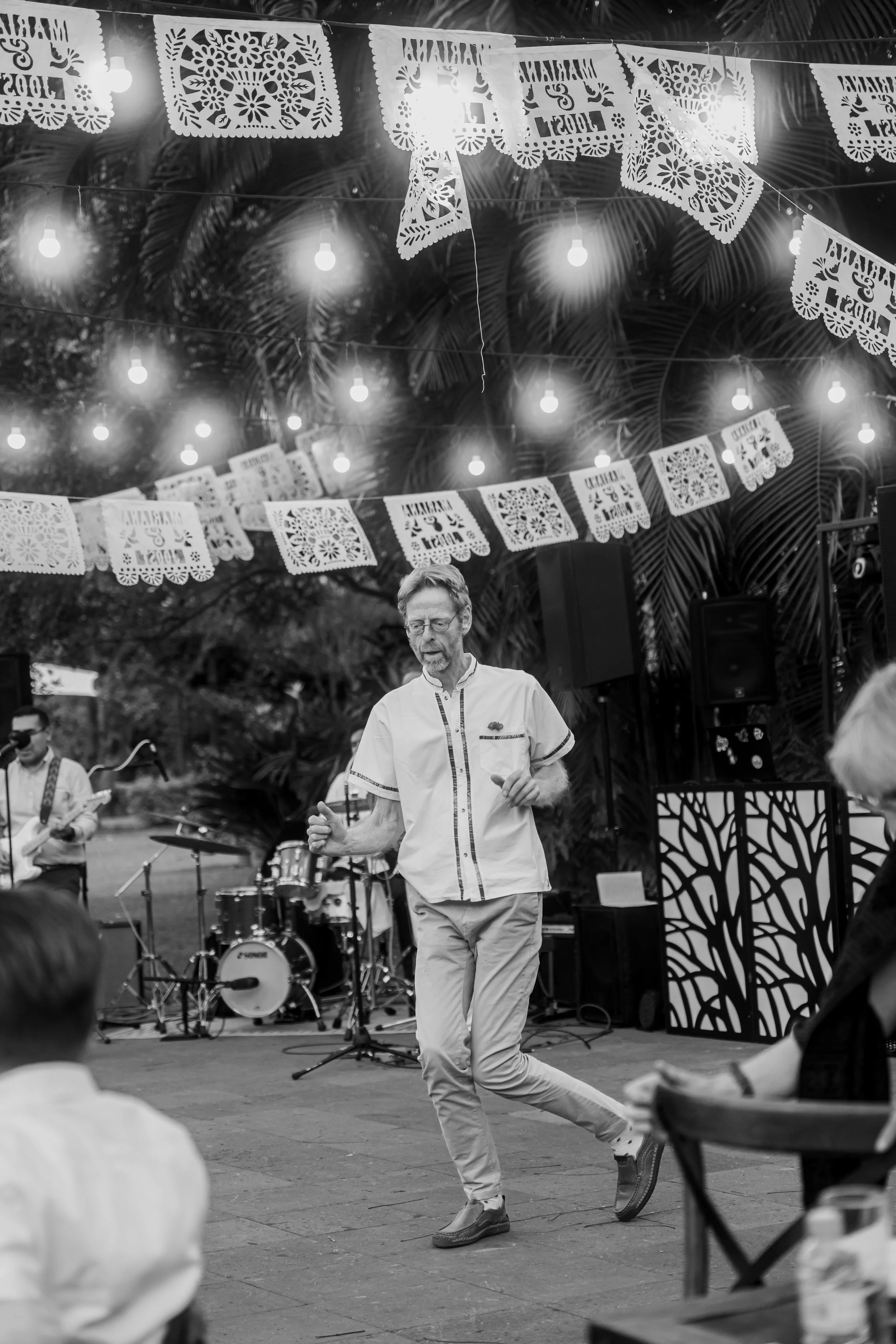 A man dancing at an outdoor celebration with papel picado banners and string lights hanging above, with a band playing in the background.