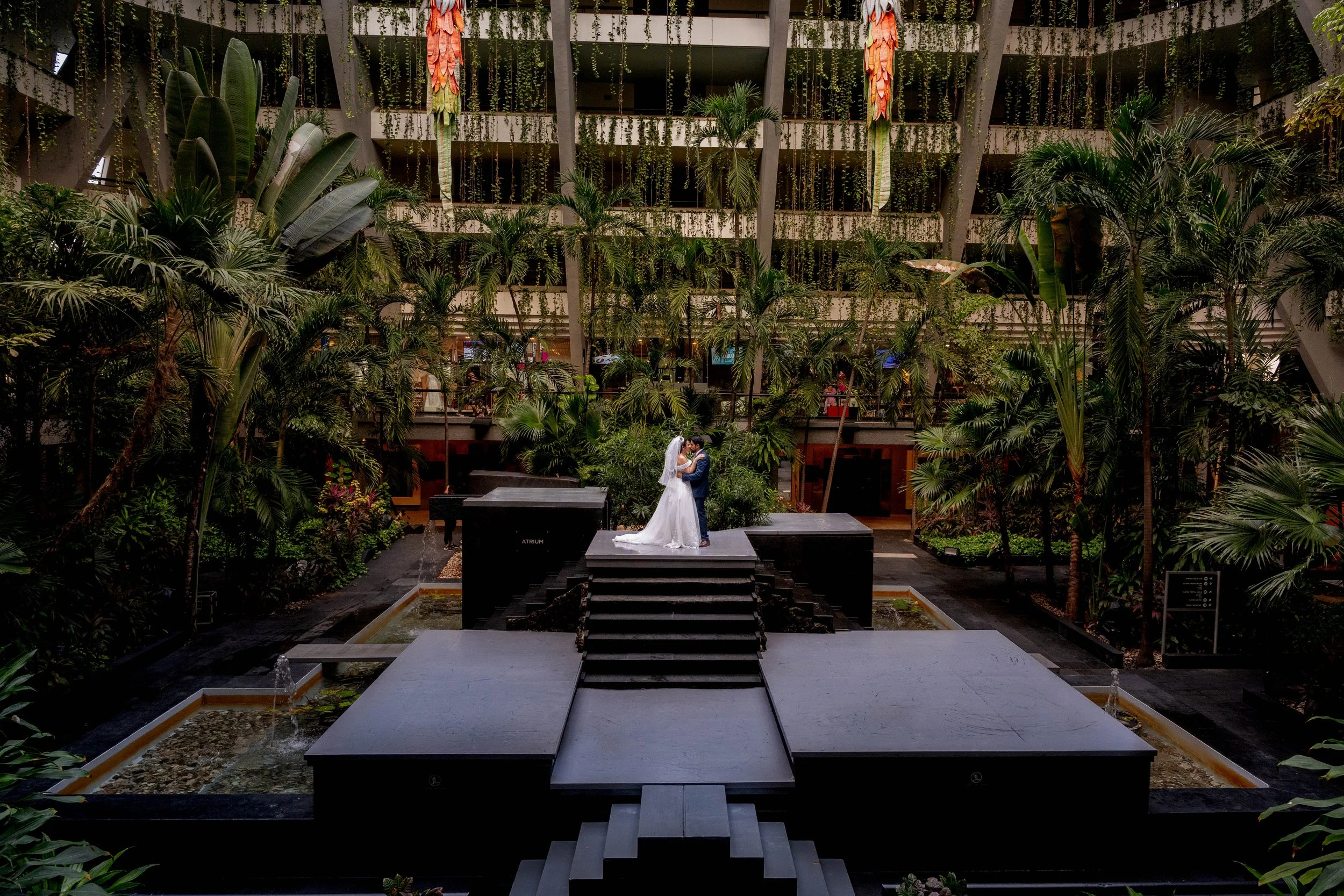 A bride and groom standing on steps in the center of an indoor lush garden, embracing and kissing under natural light, surrounded by tropical plants and trees in a hotel atrium.