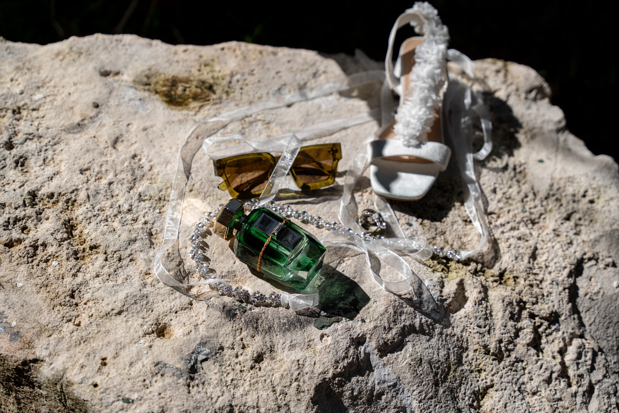 A white high-heeled sandal with frilly strap, a pair of sunglasses with yellow lenses, a green miniature perfume bottle, a silver ring, and a strand of silver beads, all placed on a large textured rock outdoors.