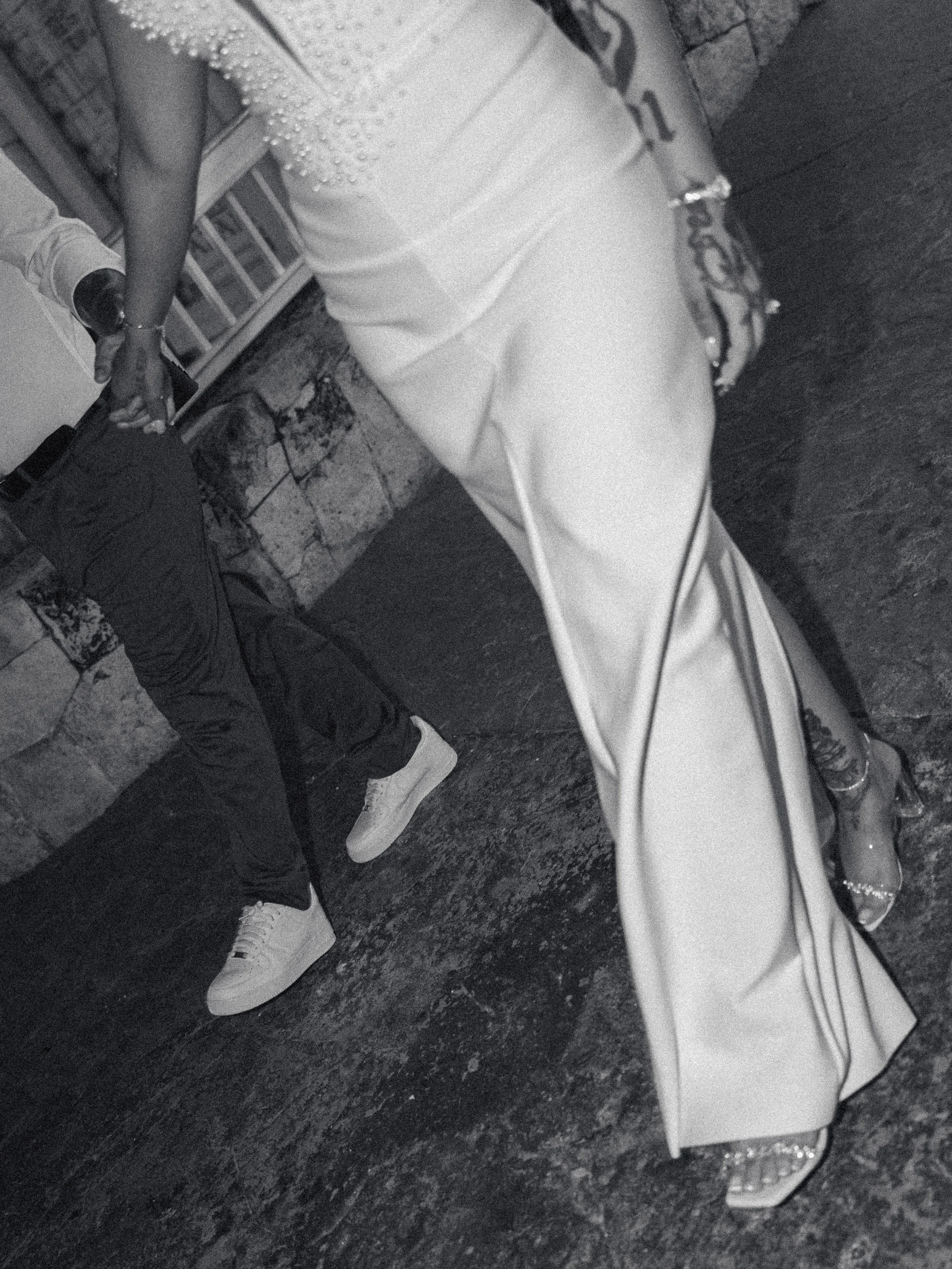 A person dressed in a white gown with embellishments, standing on a dark, textured floor, with another person in casual attire seated beside them.