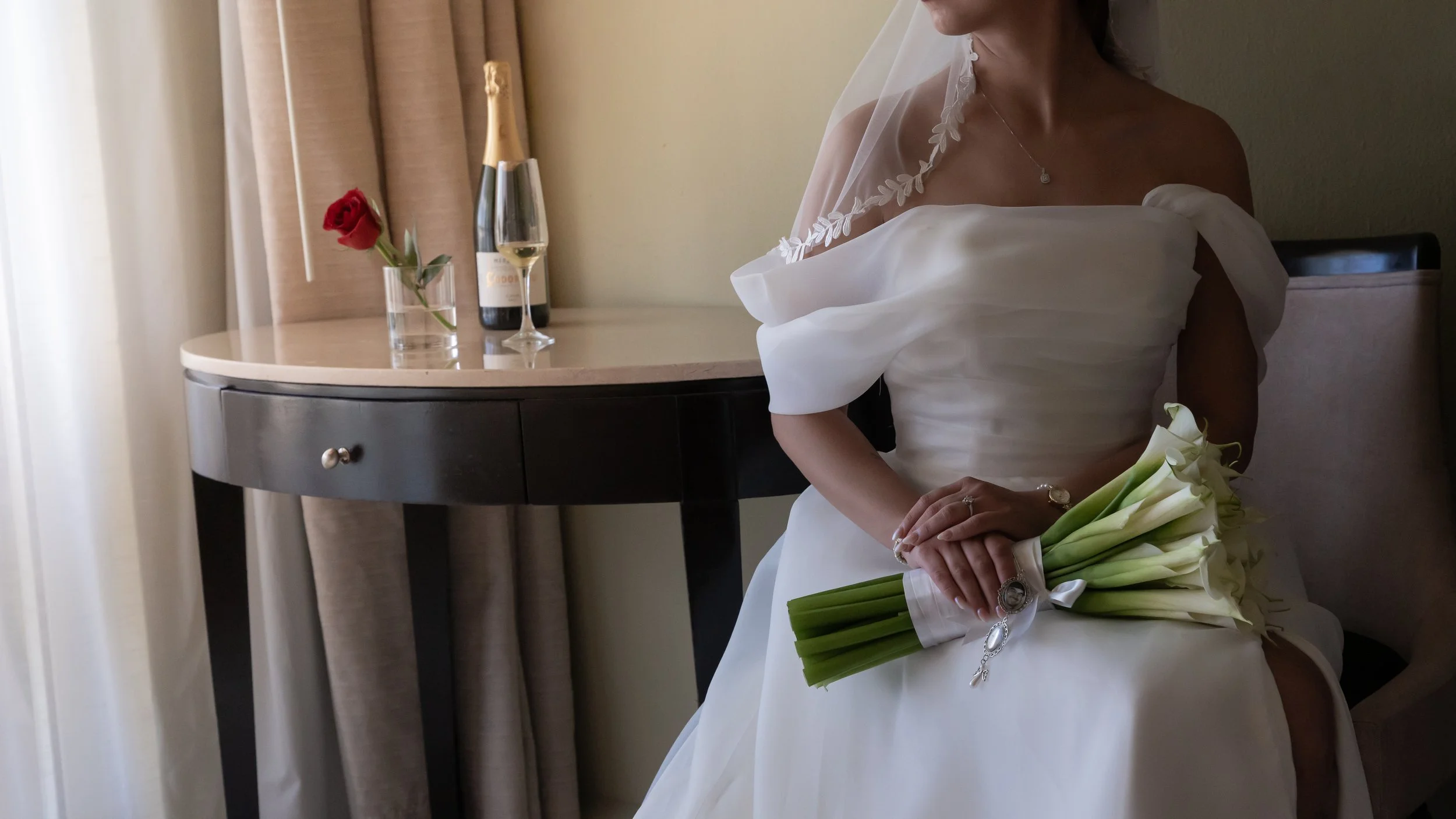 A bride sitting on a chair holding a bouquet of white calla lilies, wearing a white wedding dress and veil, with a bracelet and ring, in a room with beige curtains, a black and beige desk, a vase with a red rose, a bottle of champagne, and a glass of