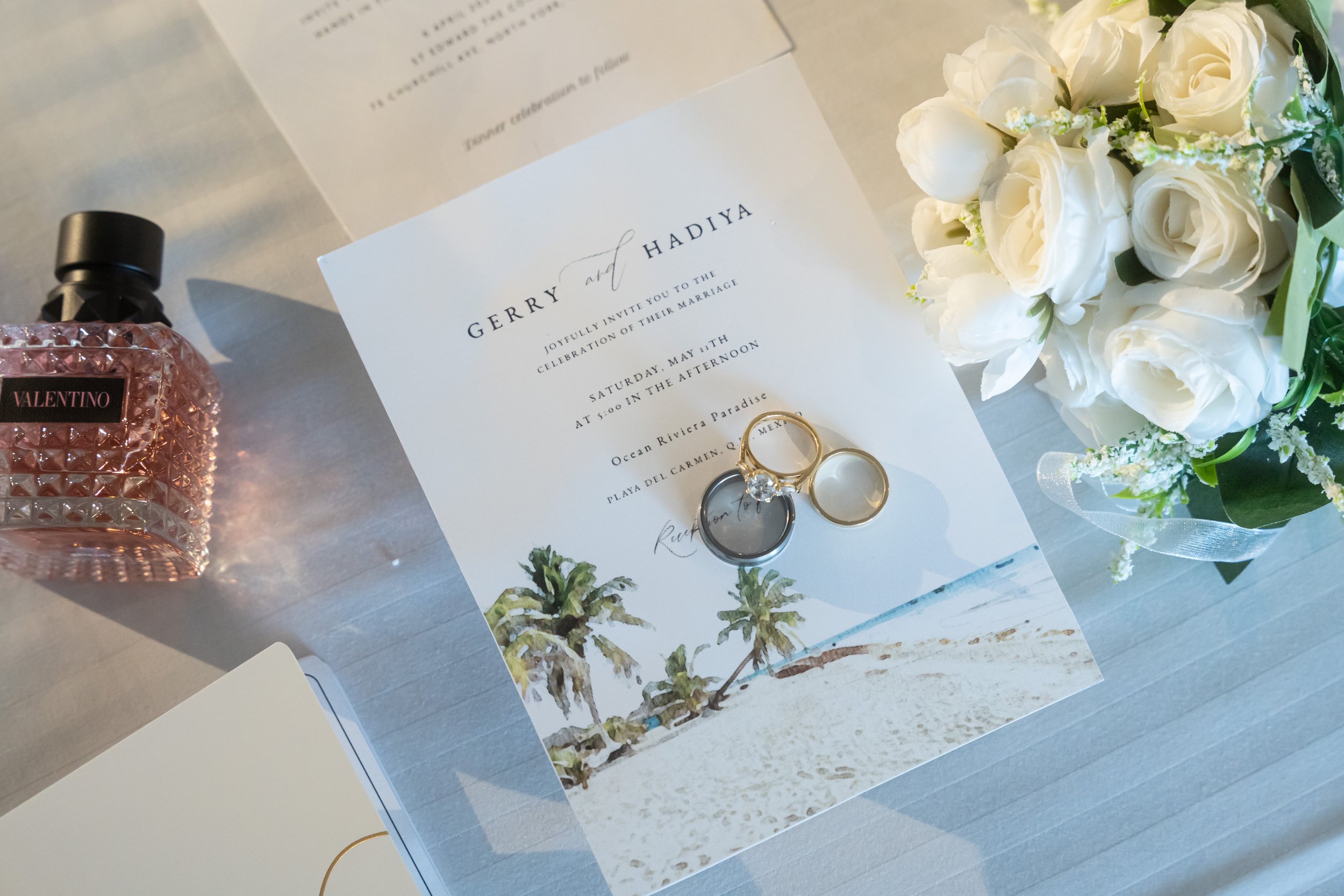 Wedding invitation card with rings on top, white bouquet of roses, Valentino perfume bottle, and part of another card visible on a white table.