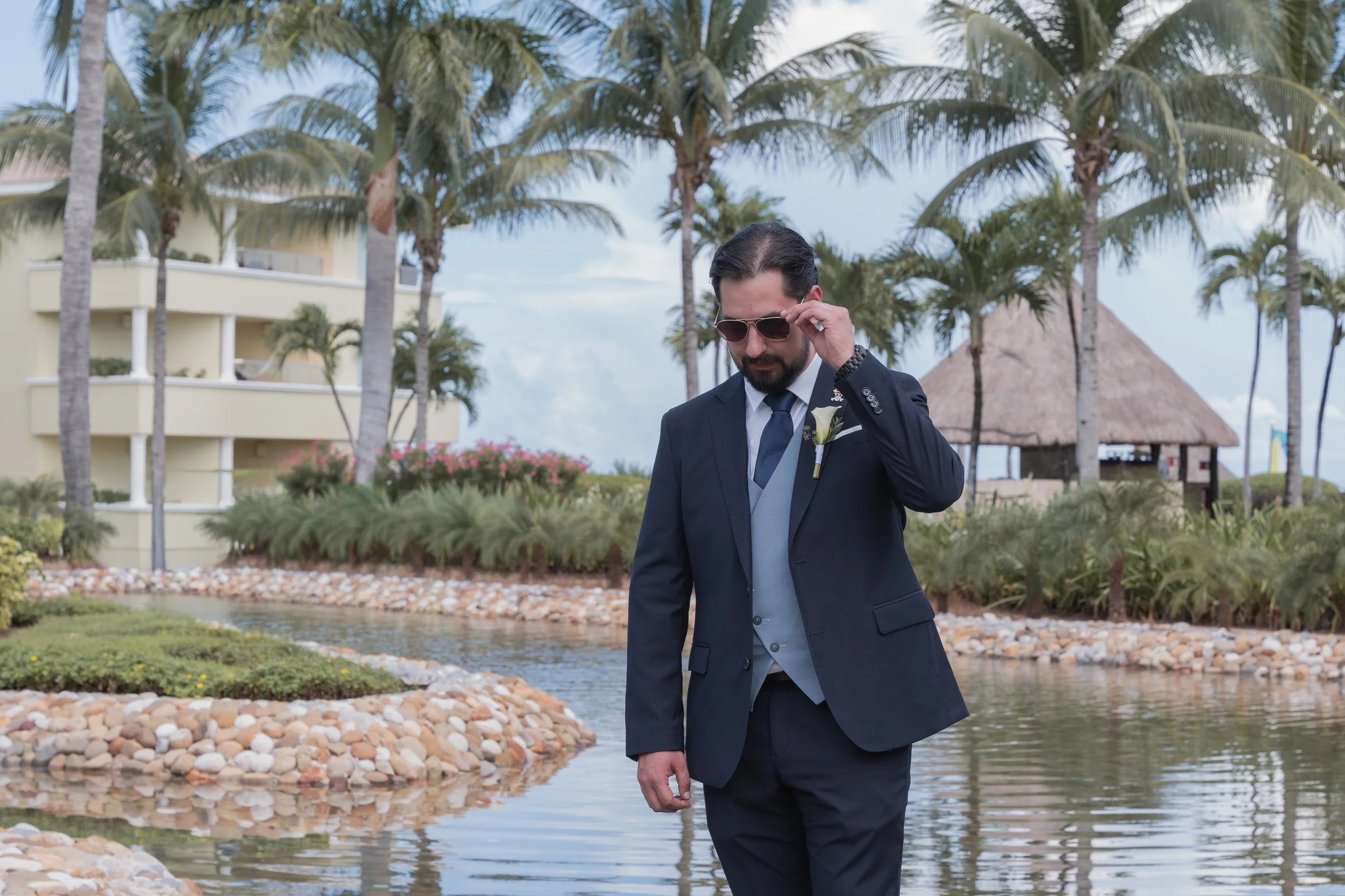 A man in a dark suit with a boutonniere, sunglasses, and a tie, standing near a water feature with rocks, tropical plants, and palm trees in the background at a beachside resort.