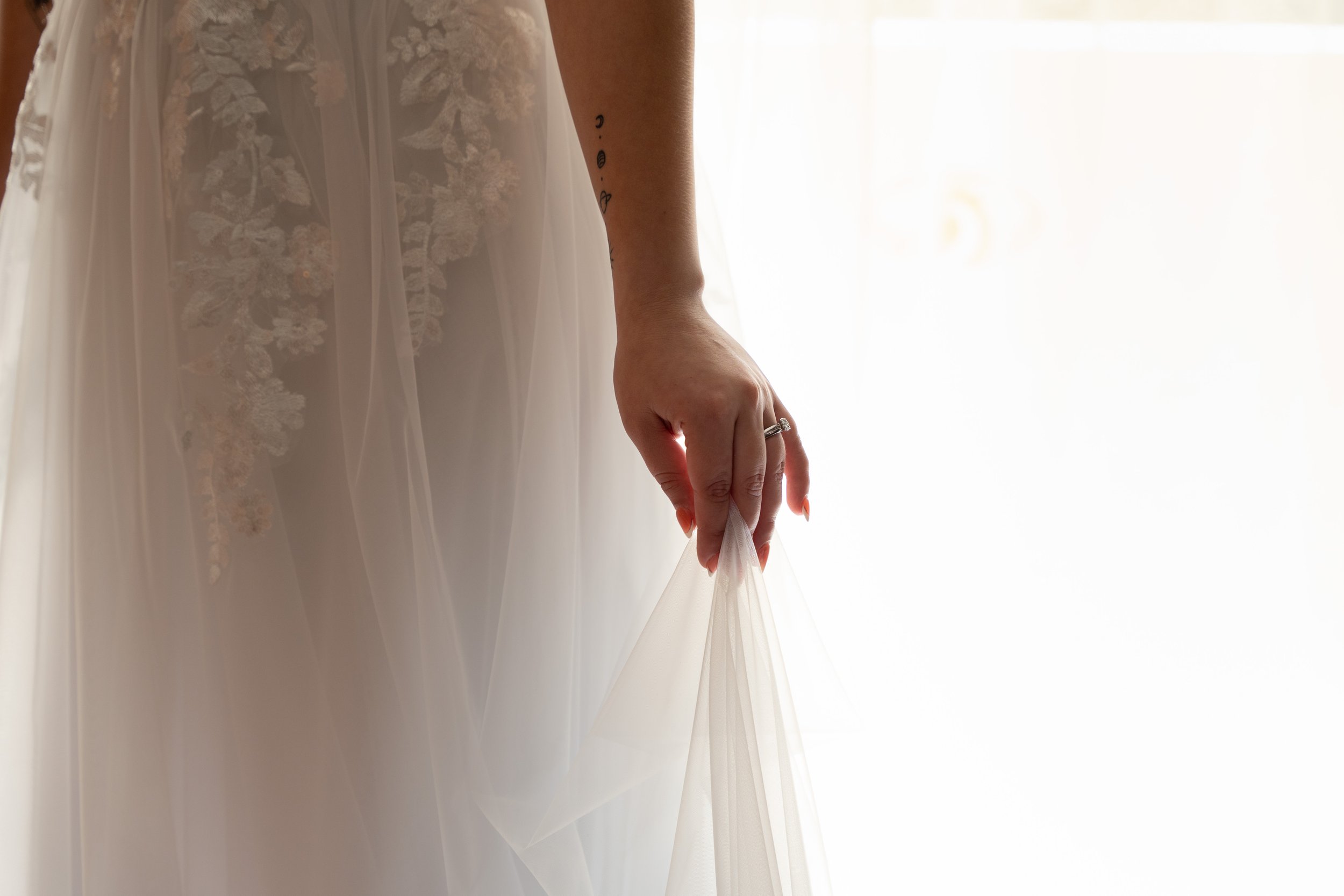 Close-up of a bride's hand holding the edge of her wedding dress in front of a bright window.