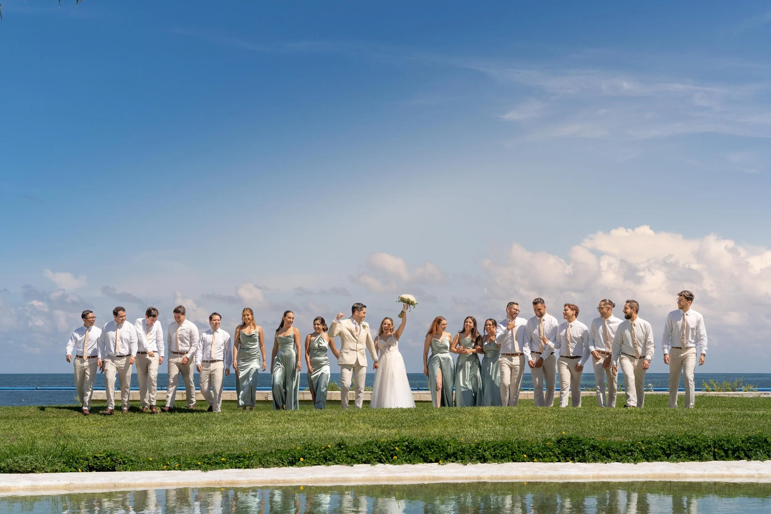 A wedding party standing outdoors on a grassy area near the ocean, with blue sky and clouds in the background. The bride and groom are in the center, with the bride holding a bouquet and wearing a white wedding dress. The bridesmaids are in matching 
