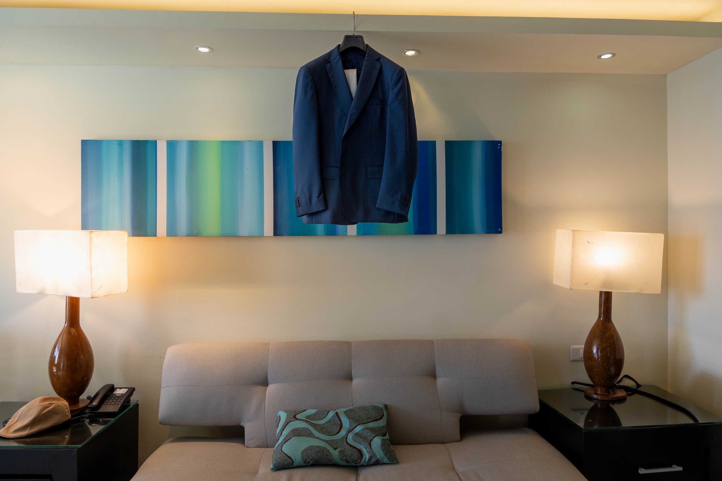 A navy blue suit jacket hanging on a black coat hanger on a wall-mounted art piece above a beige sofa with a patterned pillow. Two wooden lamps with white shades are on black side tables on either side of the sofa.