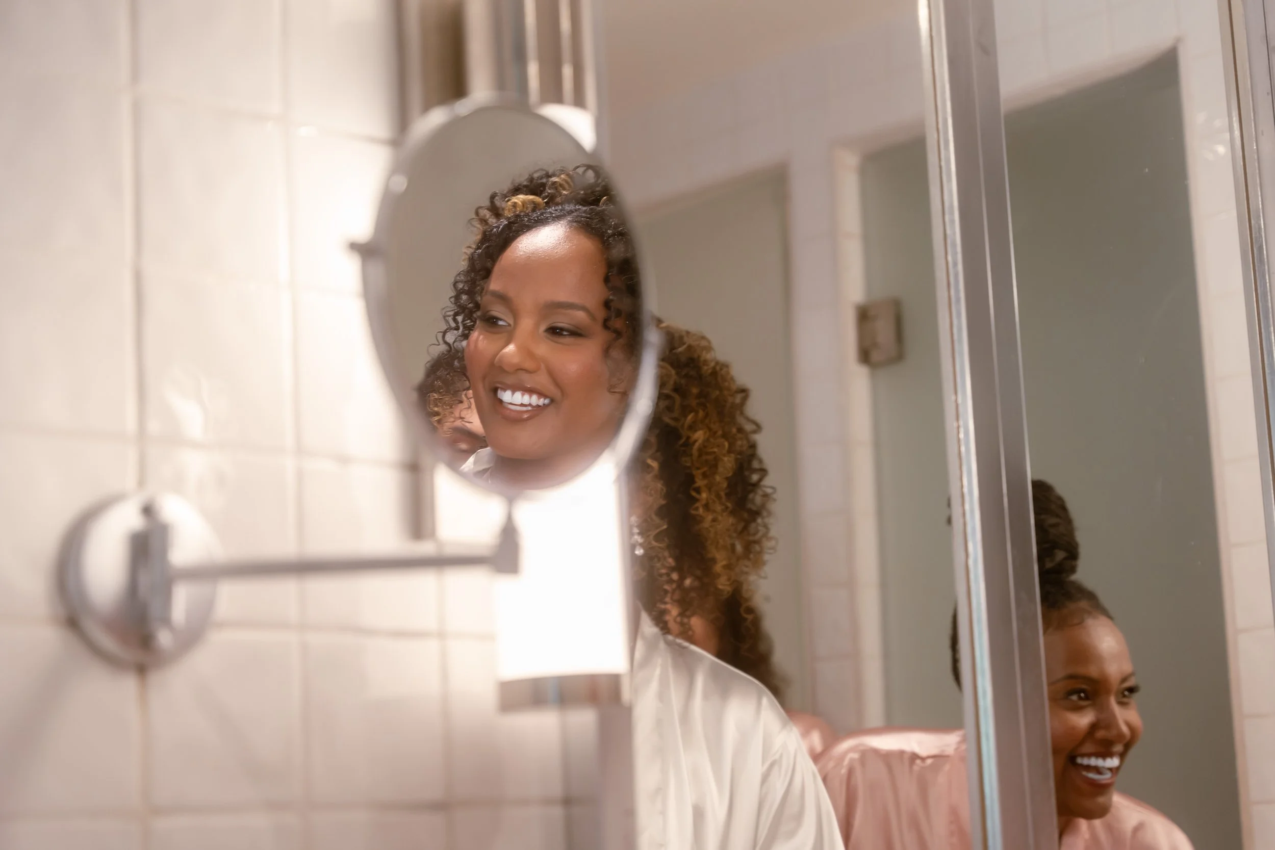 A woman with curly hair smiling while looking at her reflection in a mirror in a bathroom.