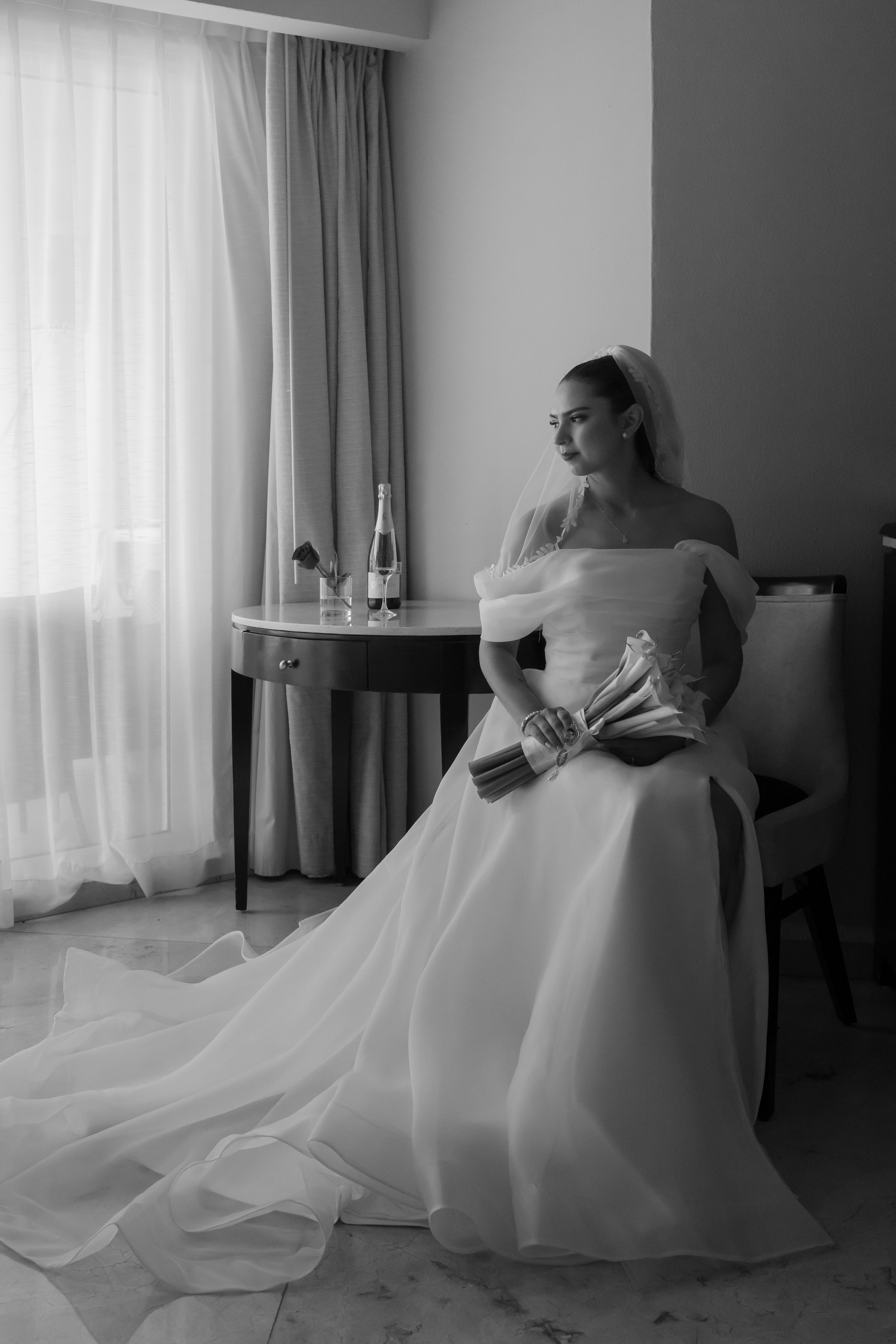 A bride sitting on a chair in a room with curtains, holding a bouquet of flowers, with a table and drinks nearby.