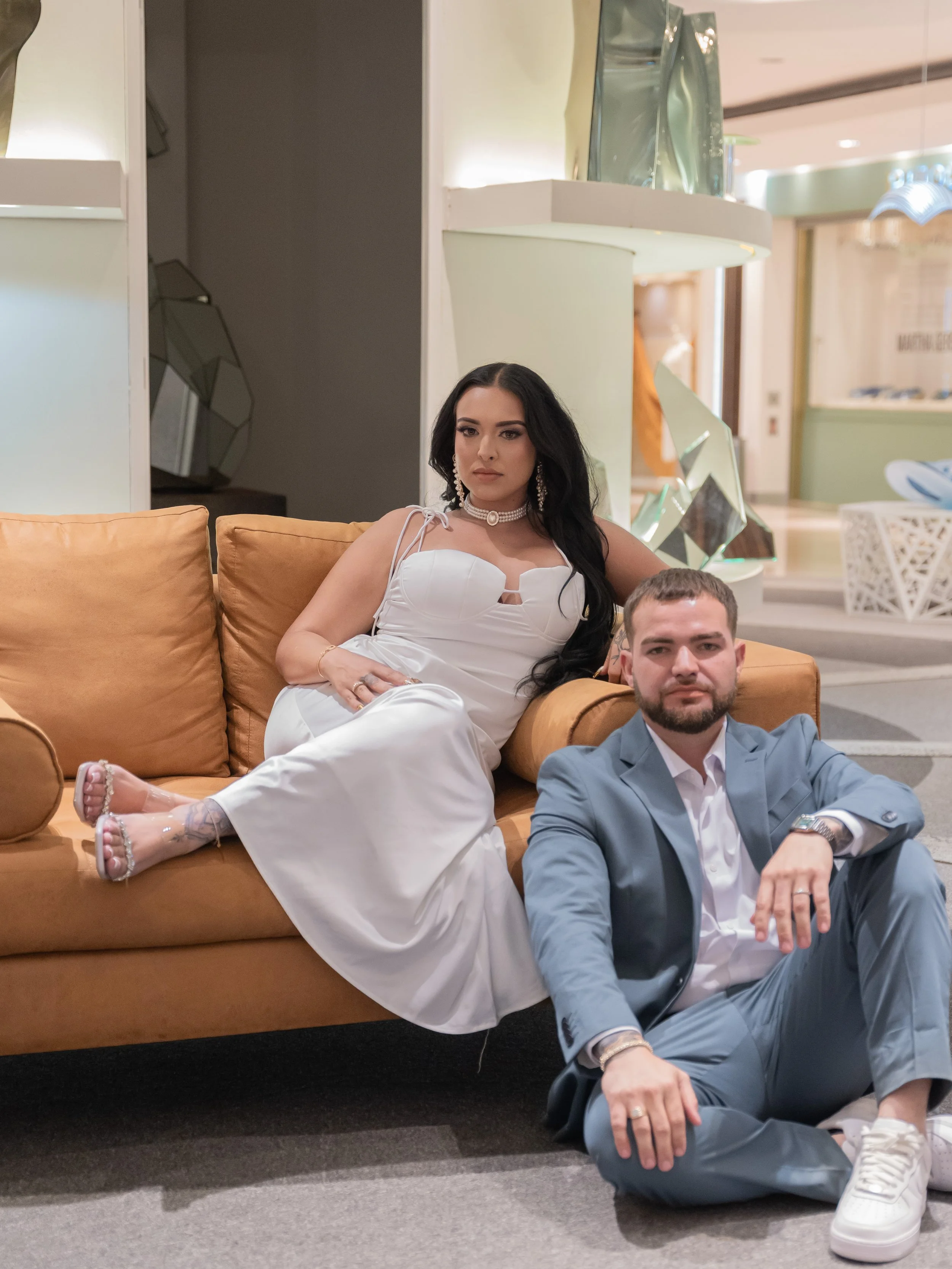 A woman and a man pose on and beside a beige couch in a modern interior space. The woman is seated comfortably on the couch, wearing a white dress and adorned with jewelry, while the man is sitting on the floor next to the couch, dressed in a light b