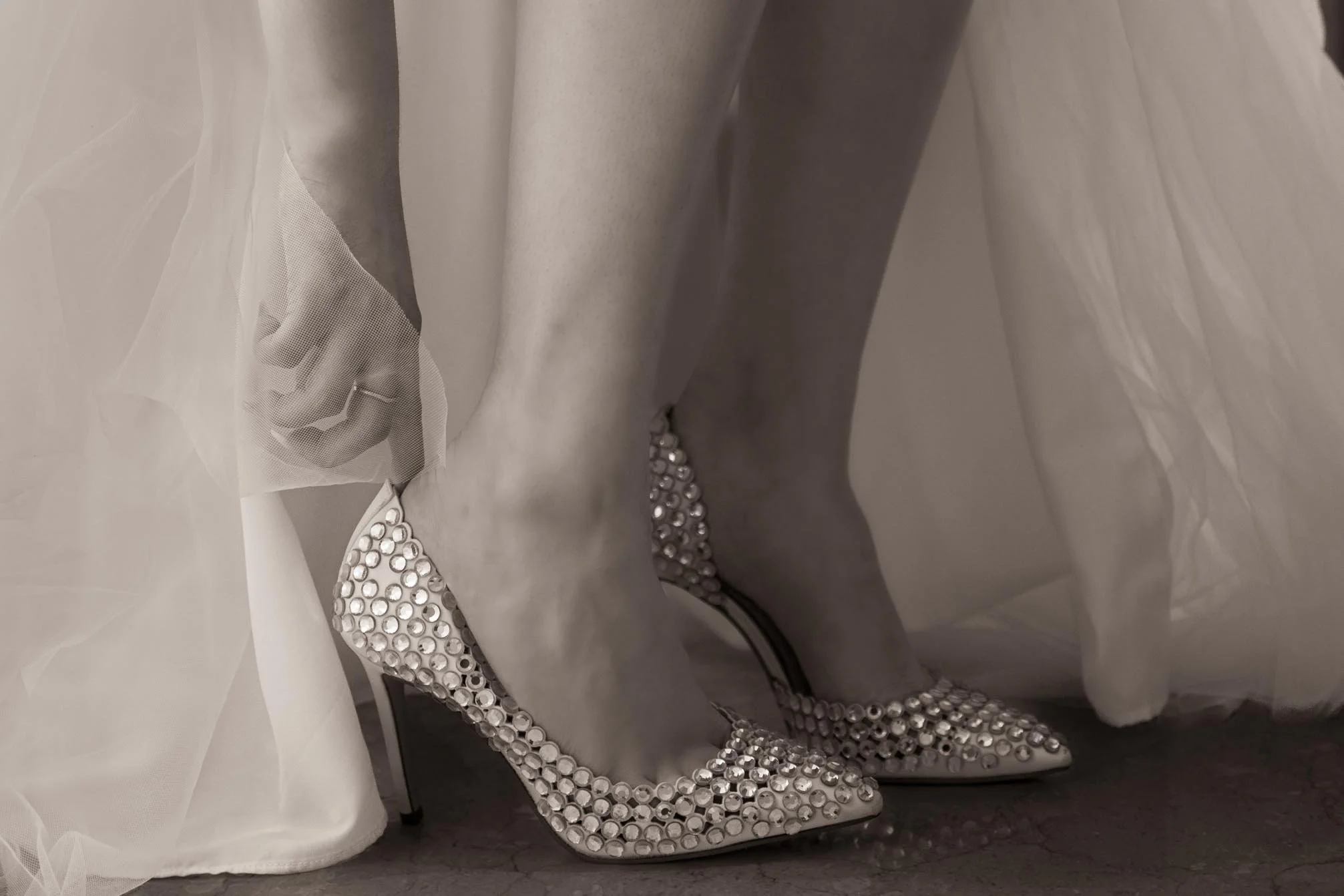 Close-up of a bride's legs and feet wearing high-heeled shoes decorated with rhinestones, with a veil partially covering her foot.