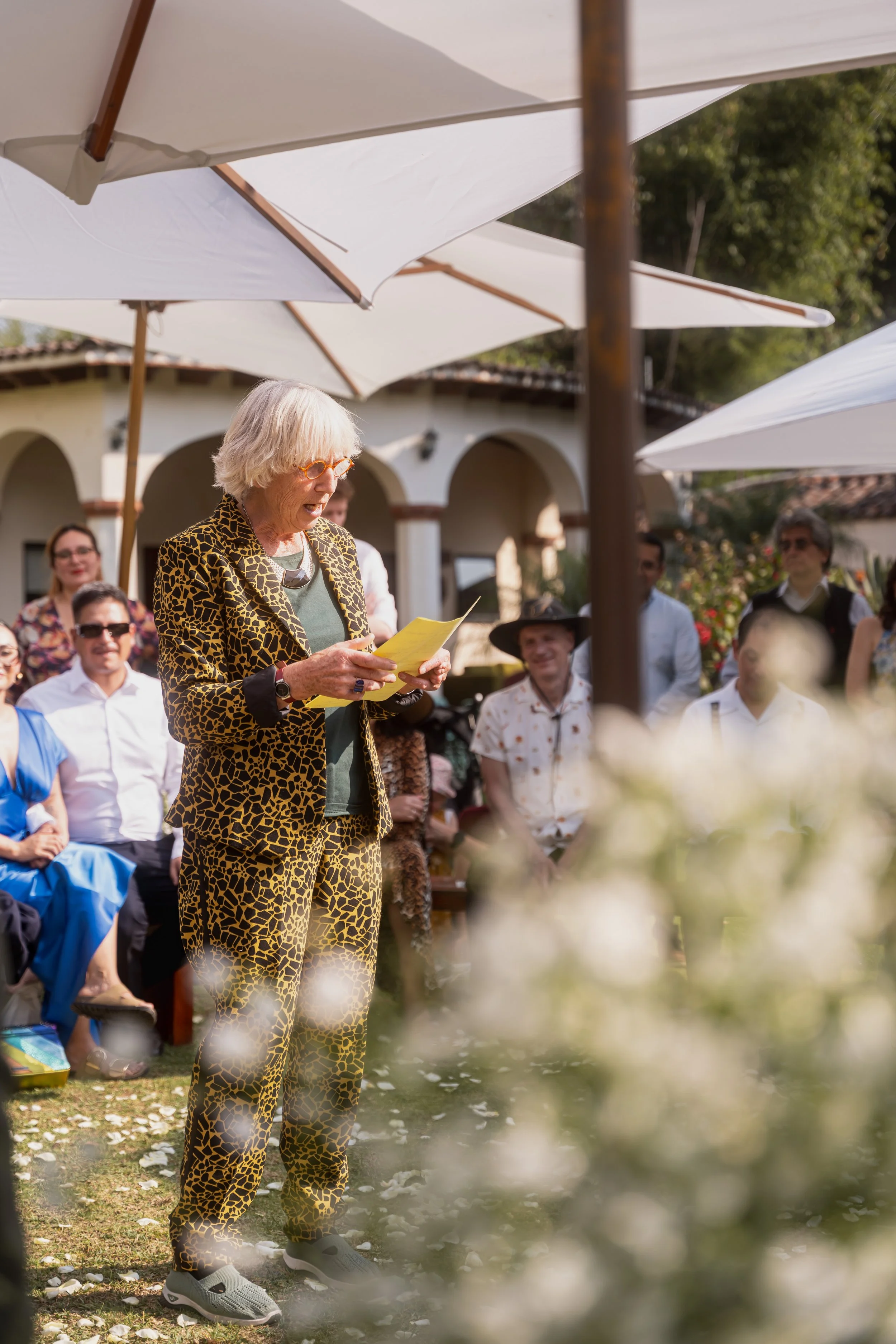A woman with white hair, wearing a leopard-print suit, reading from a yellow paper at an outdoor gathering under white umbrellas. Several people seated and standing behind her, some smiling, in front of a building with arches and greenery.