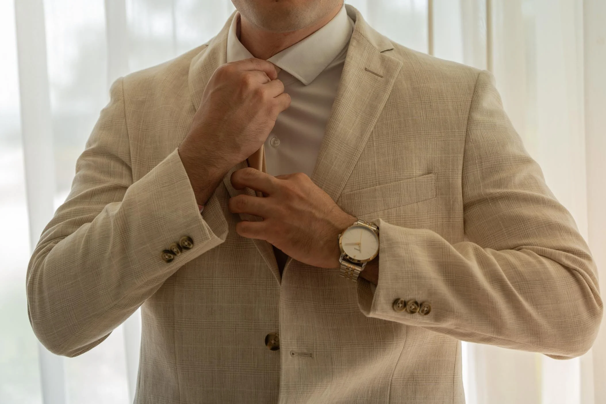A man in a cream-colored suit adjusting his tie, wearing a white dress shirt and a gold wristwatch.