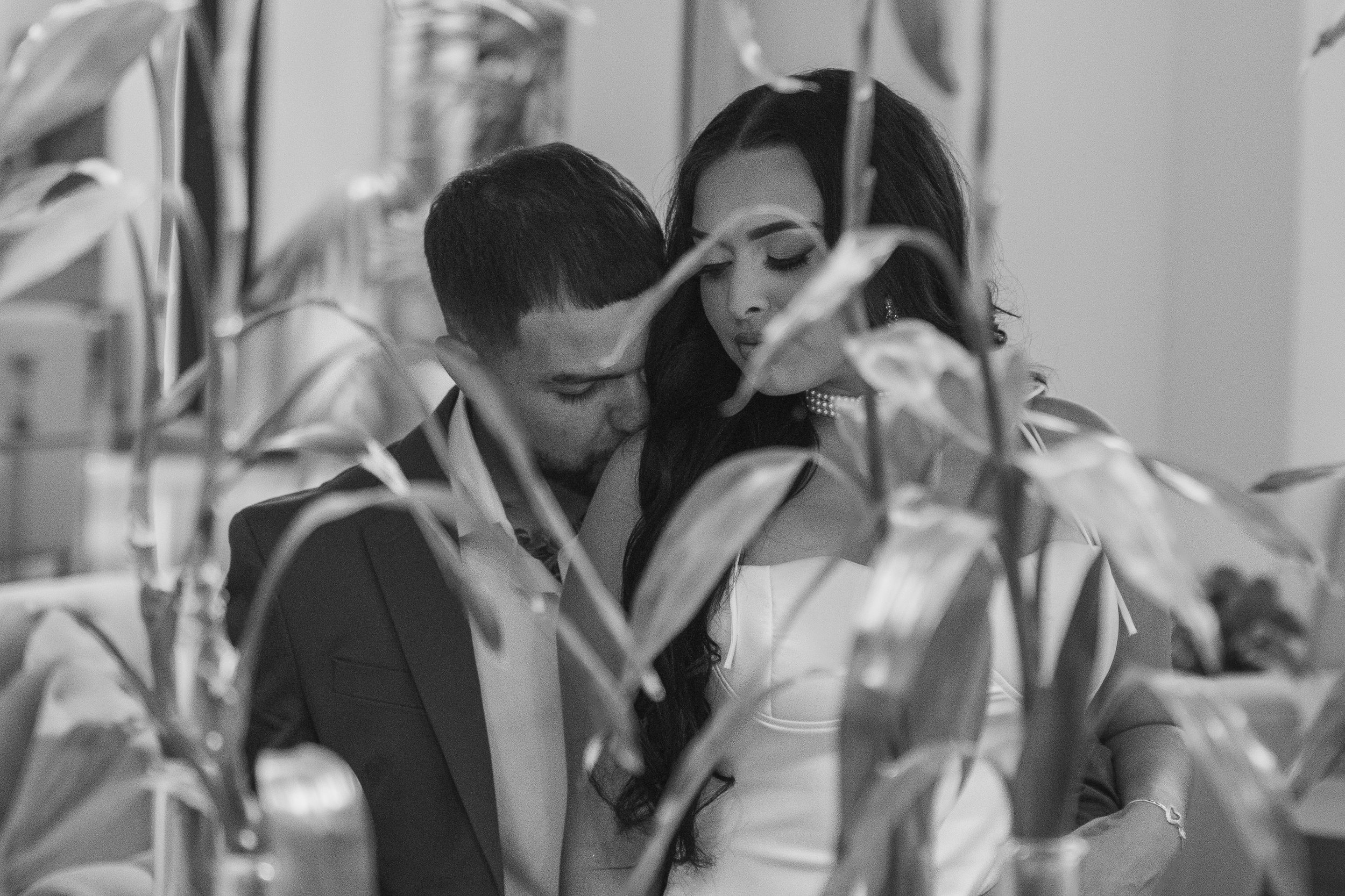 A black and white photo of a couple embracing closely indoors, framed by large plant leaves in the foreground.