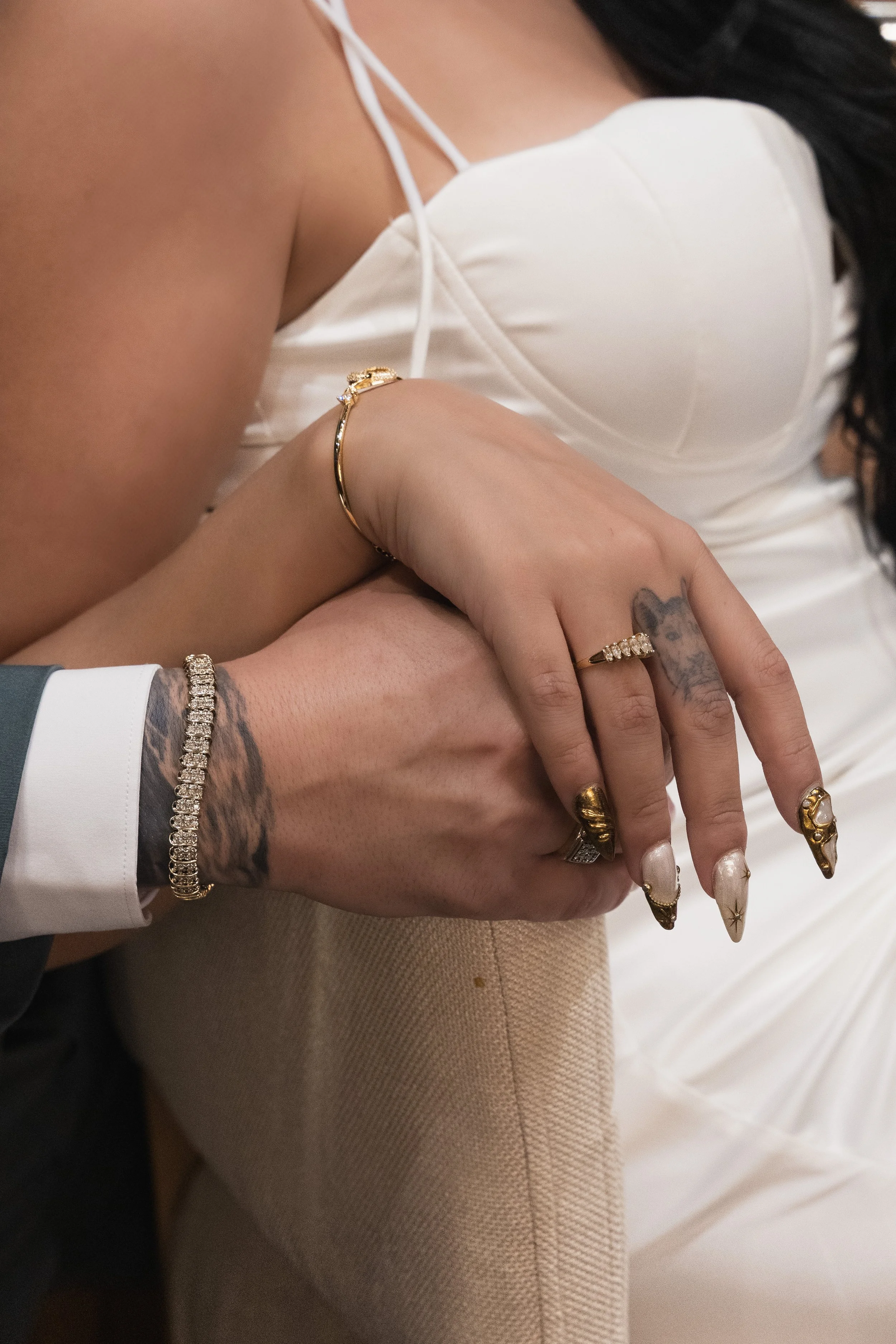 Close-up of two hands intertwined, showing jewelry and tattoos against a background of the woman's white satin dress.