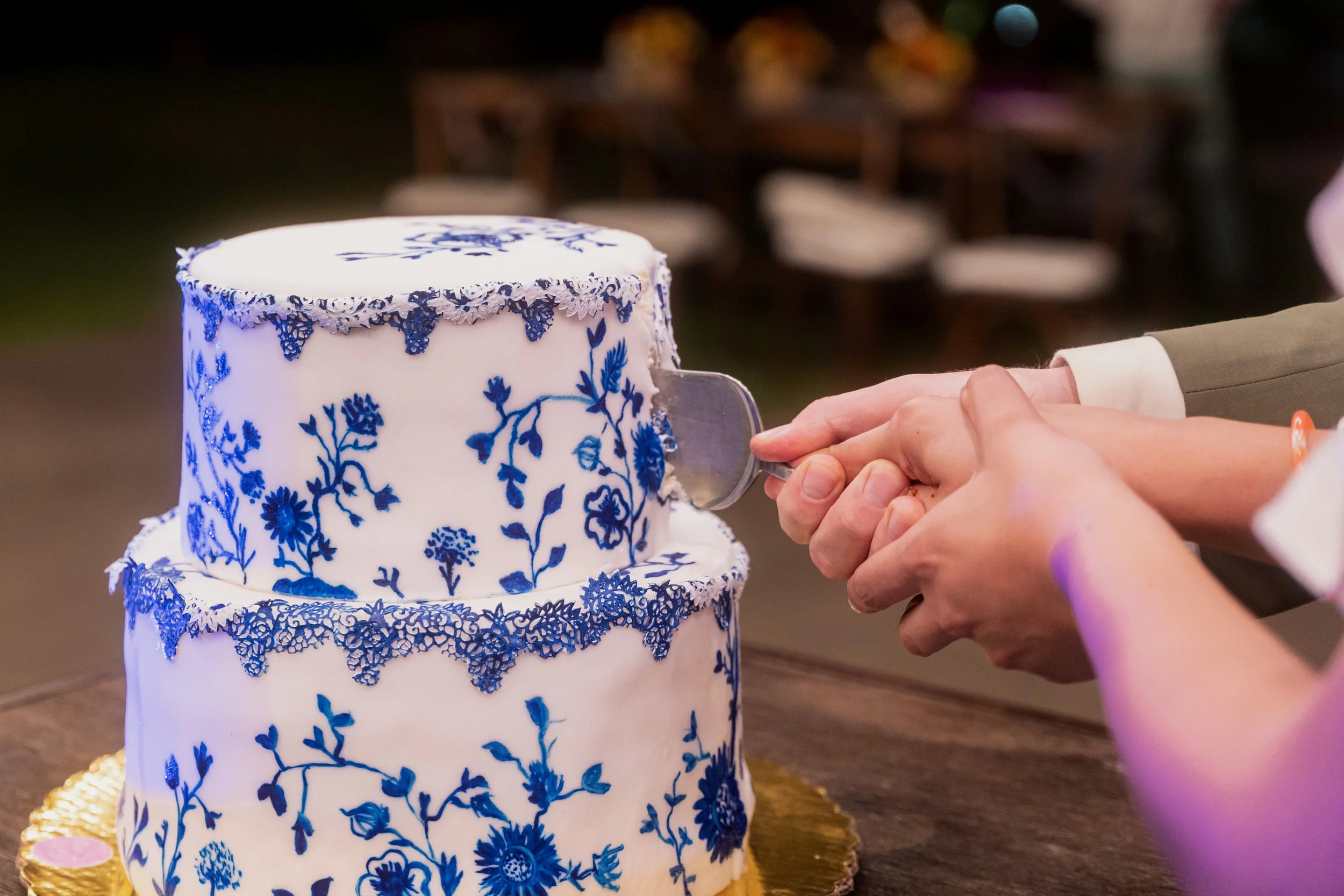 A three-tiered wedding cake decorated with blue floral patterns and lace-like adornments, being cut by a bride and groom's hands.