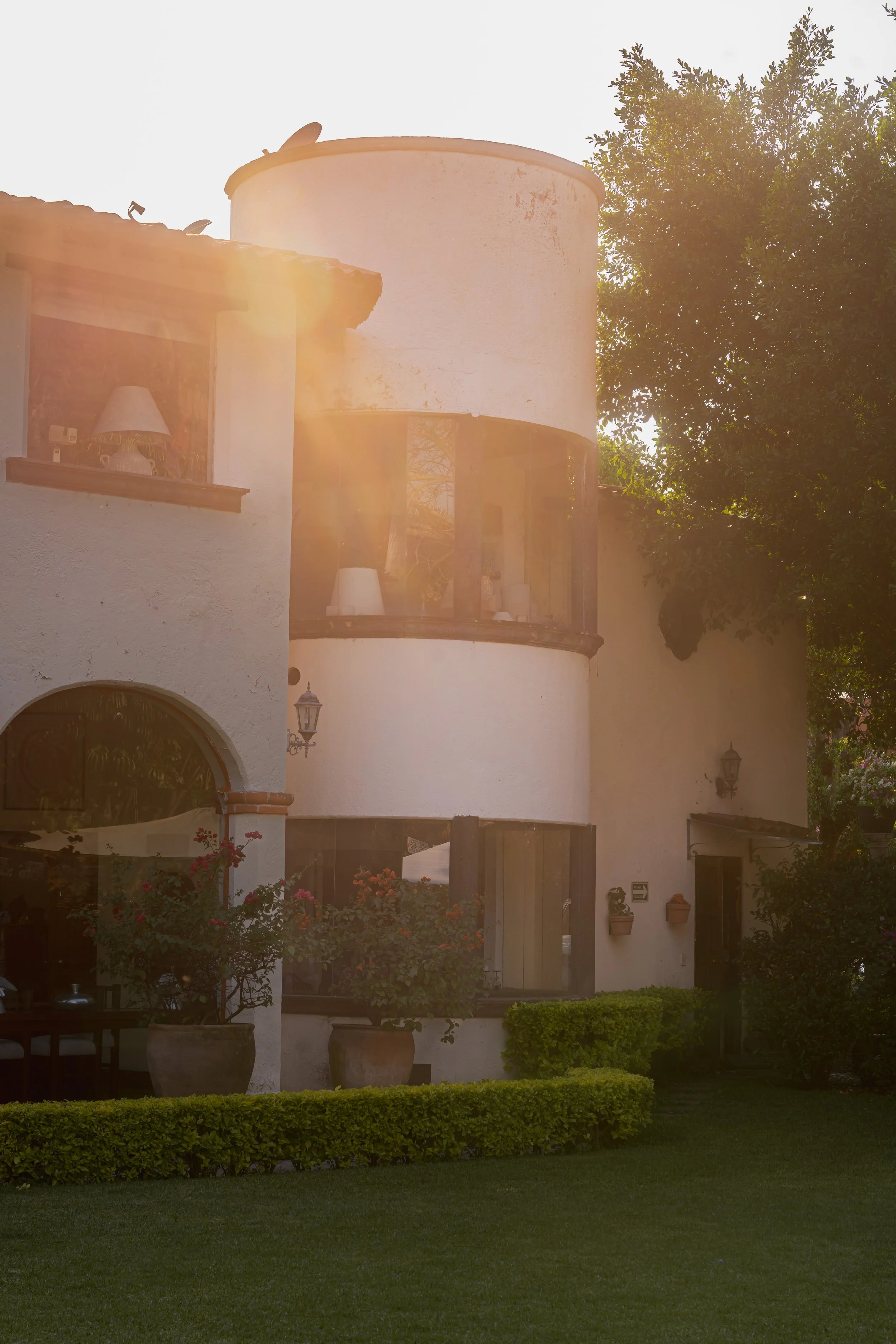 A Mediterranean-style house with white stucco walls, a rounded tower with large windows, potted plants, and a lush green lawn, bathed in sunlight.