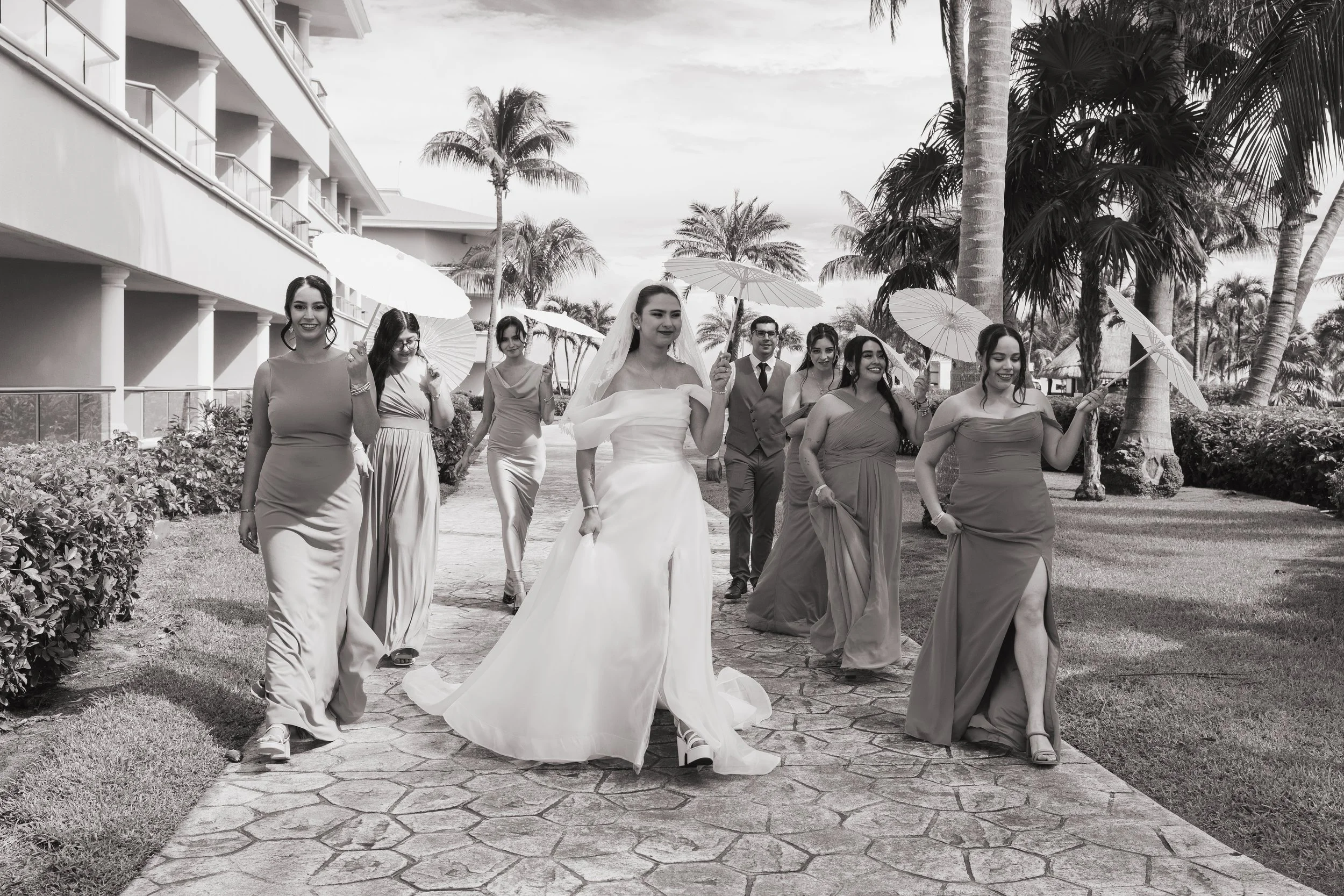 A group of women and a man walking on a stone pathway outdoors, with palm trees and a building in the background, some women holding umbrellas, one woman in wedding dress leading the group.