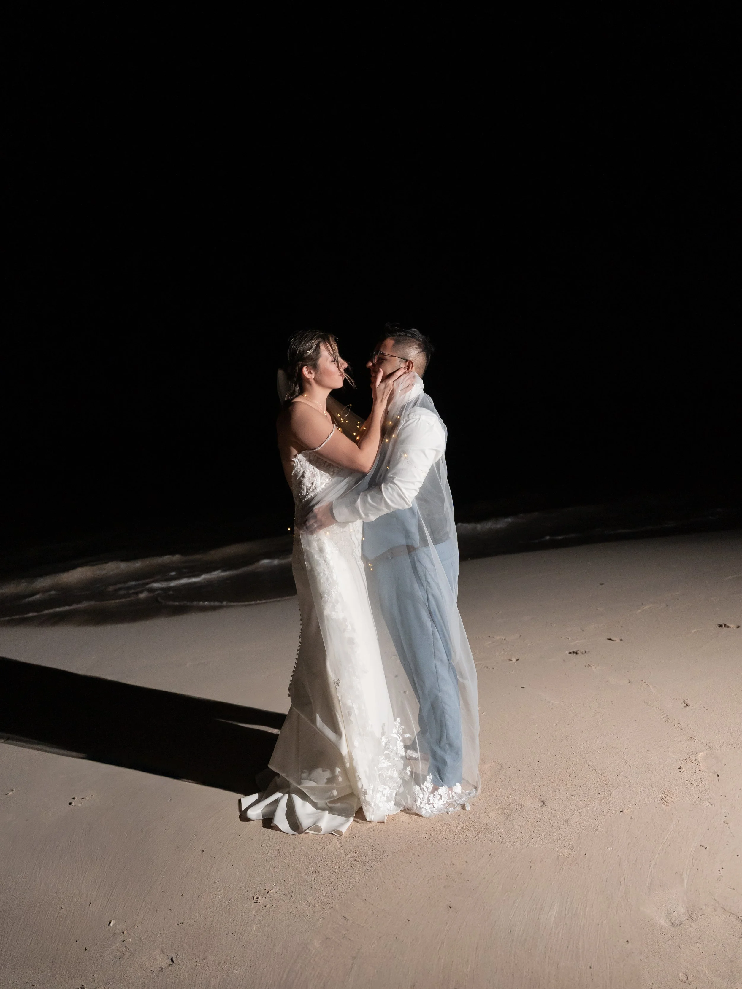 A bride and groom in wedding attire embracing at night on a beach, with the ocean in the background and a dark sky overhead.