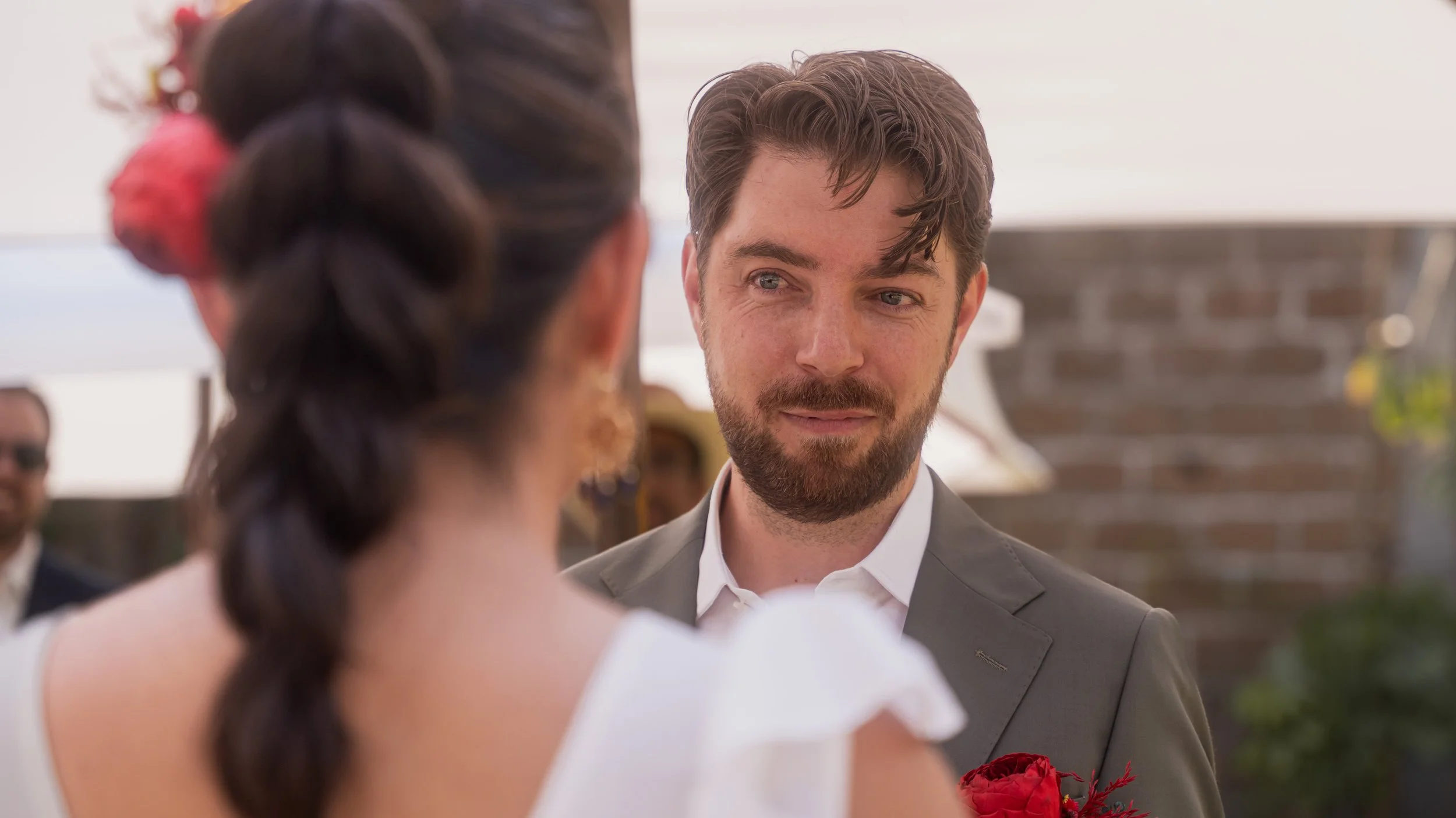 A man and woman at a wedding ceremony outdoors. The man, with a beard and wavy hair, is looking at the woman, who has dark hair styled in braids with a pink hair accessory. The woman holds a bouquet of red flowers, and there are guests and a brick wa