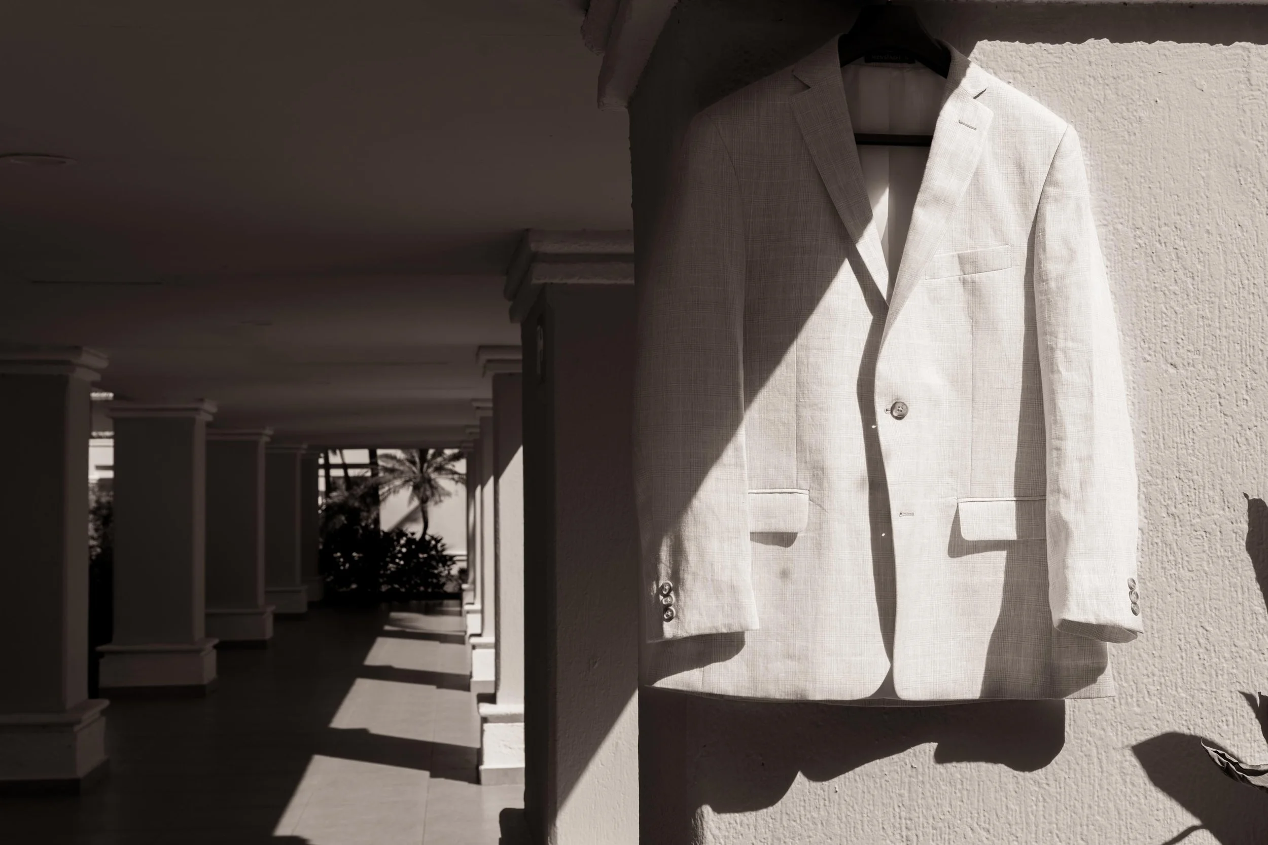 A white blazer hanging on a black hanger on a textured white wall, partially lit by sunlight with shadows cast on the wall and blazer, with a corridor and palm trees visible in the background.