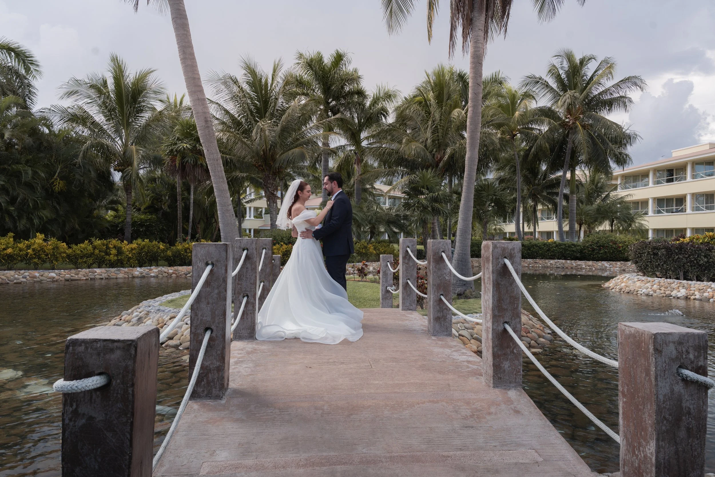 Bride and groom dancing on a small wooden bridge over water, surrounded by palm trees and tropical greenery, with resort buildings in the background.