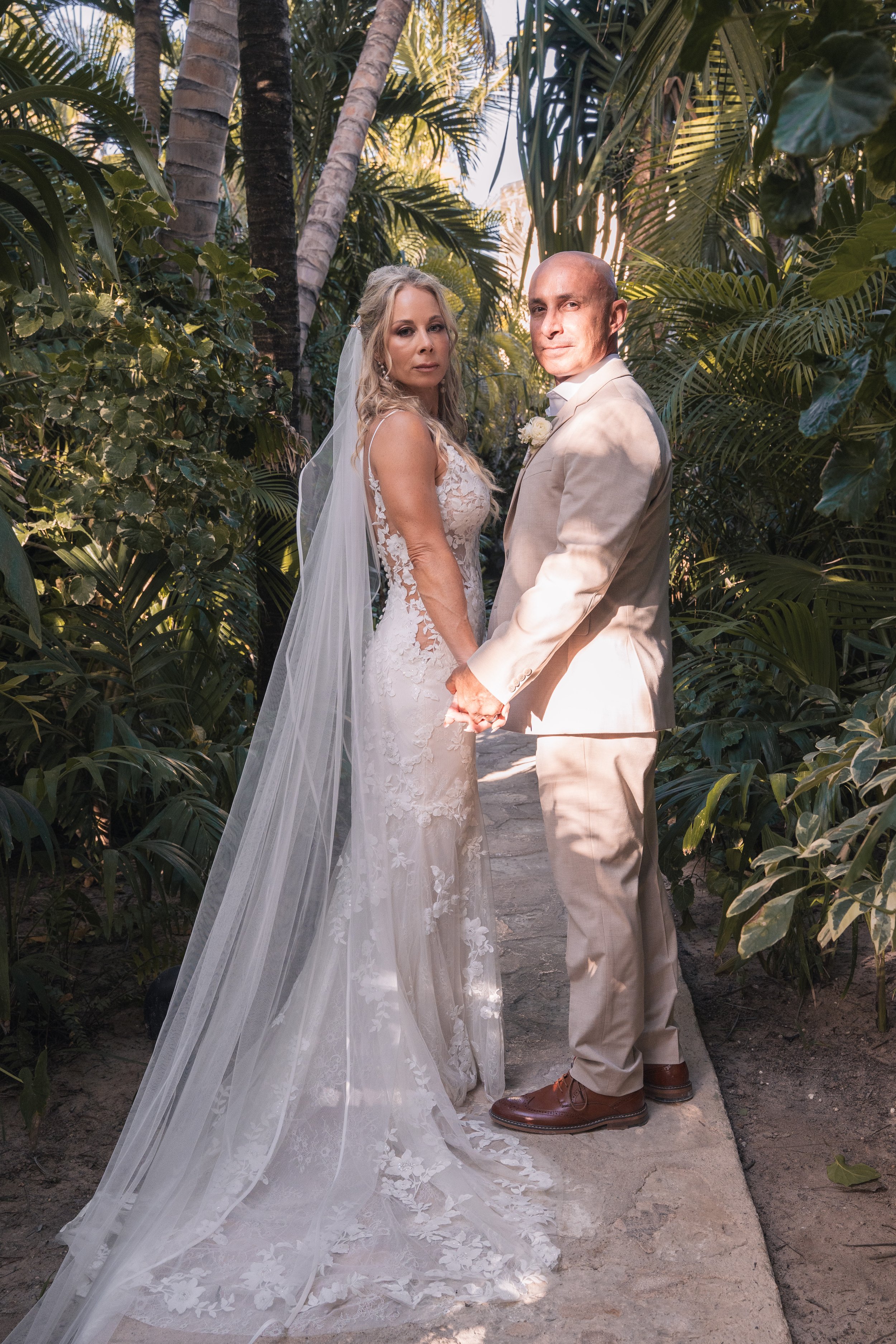 A bride and groom holding hands outdoors on a narrow stone pathway surrounded by lush green tropical plants, with sunlight filtering through the trees.