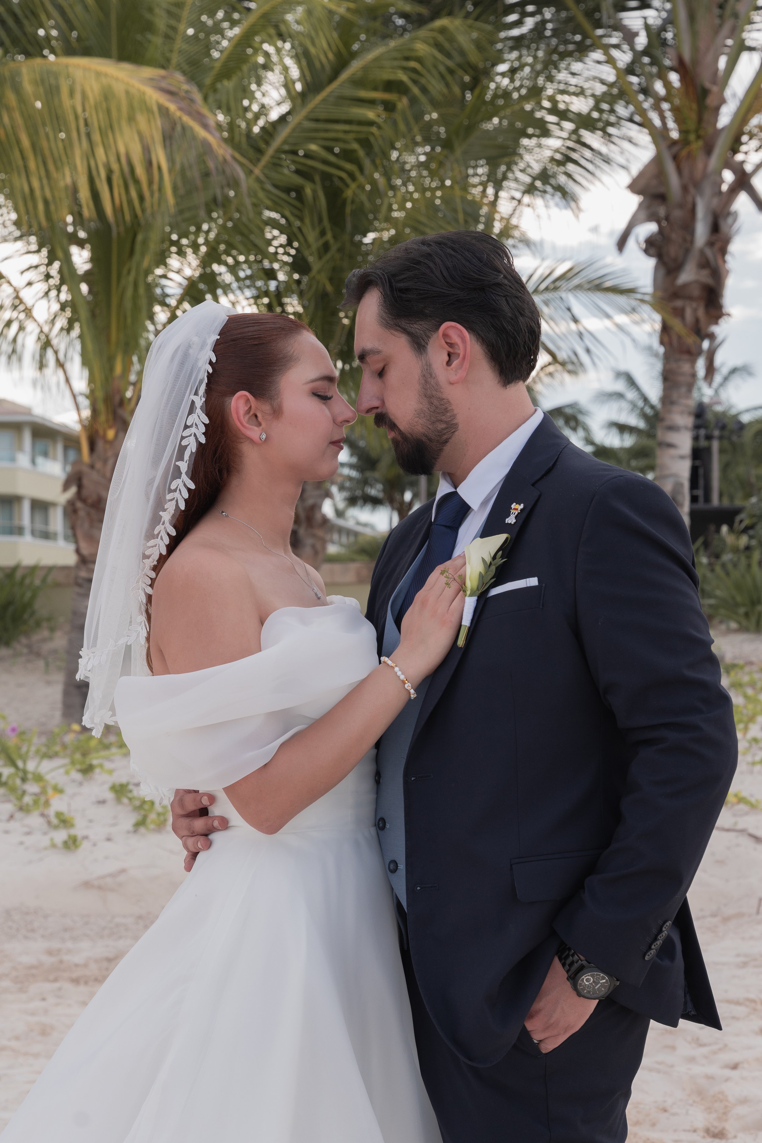 Bride and groom standing close together on a beach, with palm trees in the background, their foreheads touching in a romantic moment.
