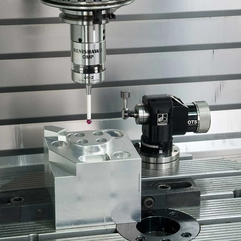 Close-up of a CMM milling machine tool performing drilling or milling on a metal block, with precision fixtures and equipment visible.
