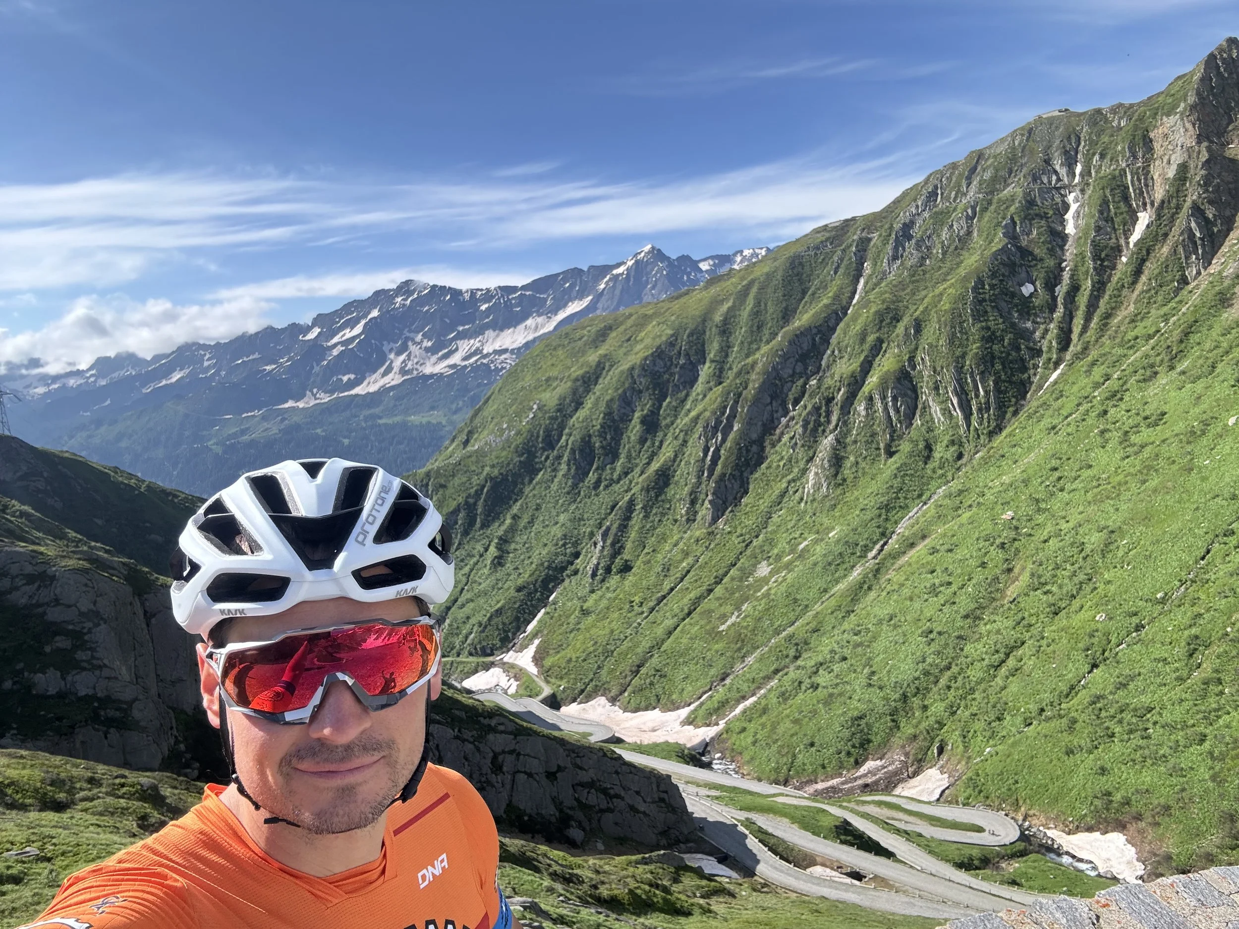 A man in cycling gear, including a white helmet and red sunglasses, taking a selfie in a mountainous area with green hills, winding roads, and snow-capped peaks in the background.