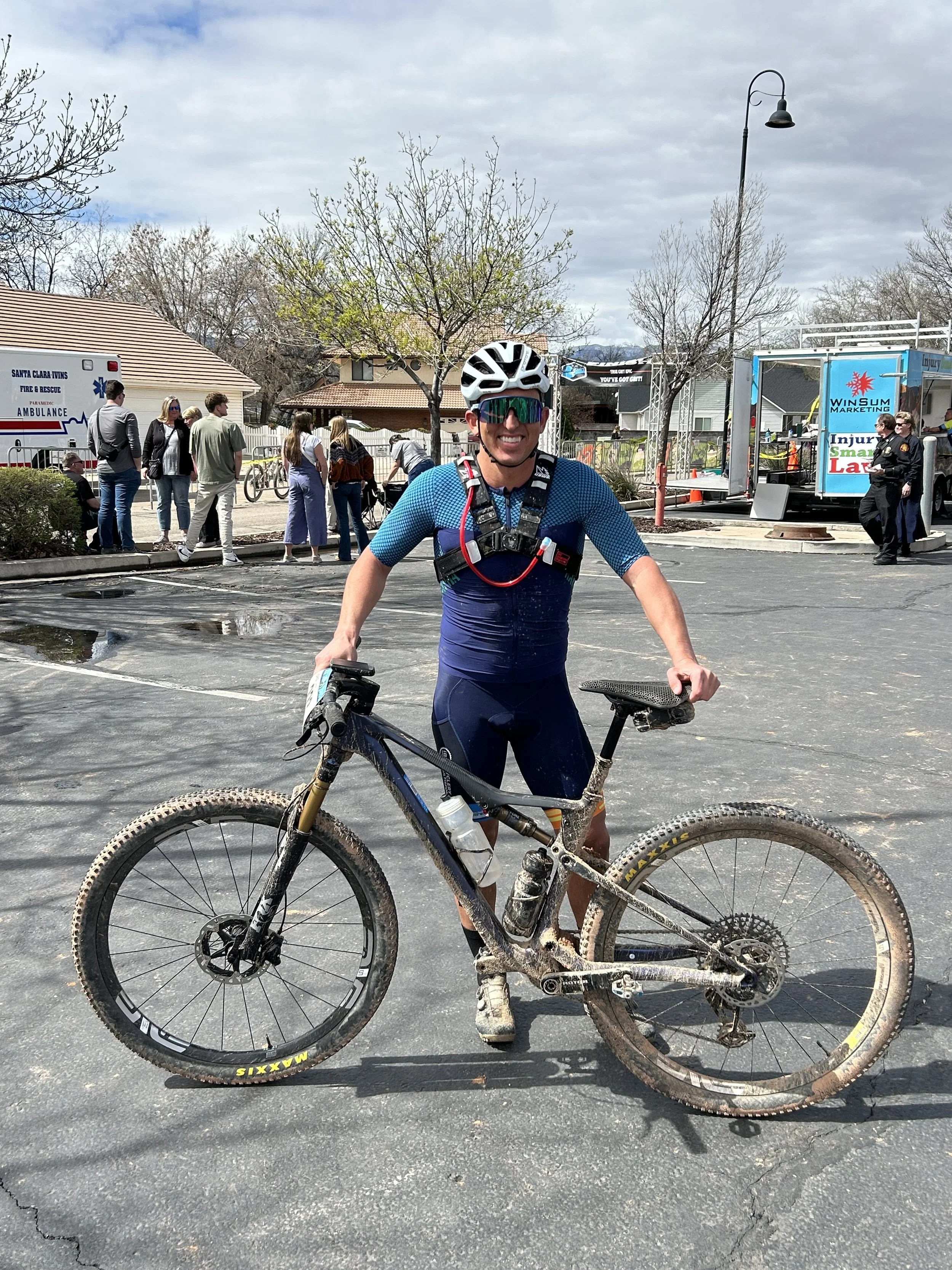 A cyclist standing next to a muddy mountain bike, smiling, wearing a helmet, sunglasses, and cycling gear in a parking lot after a ride.