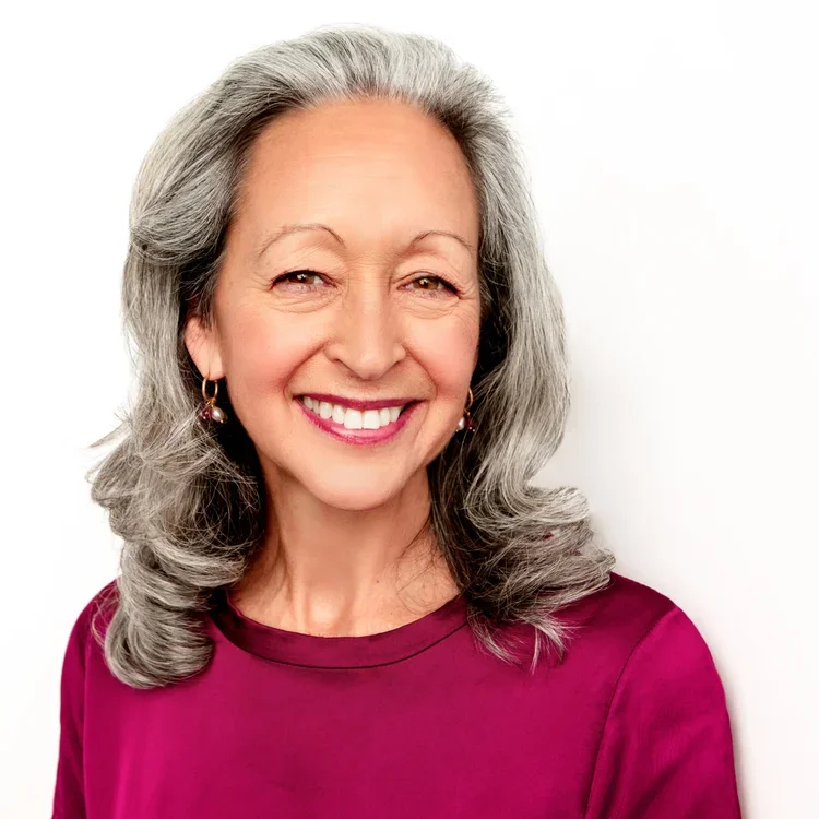 An elderly woman with long gray hair smiling and wearing a magenta top and earrings.