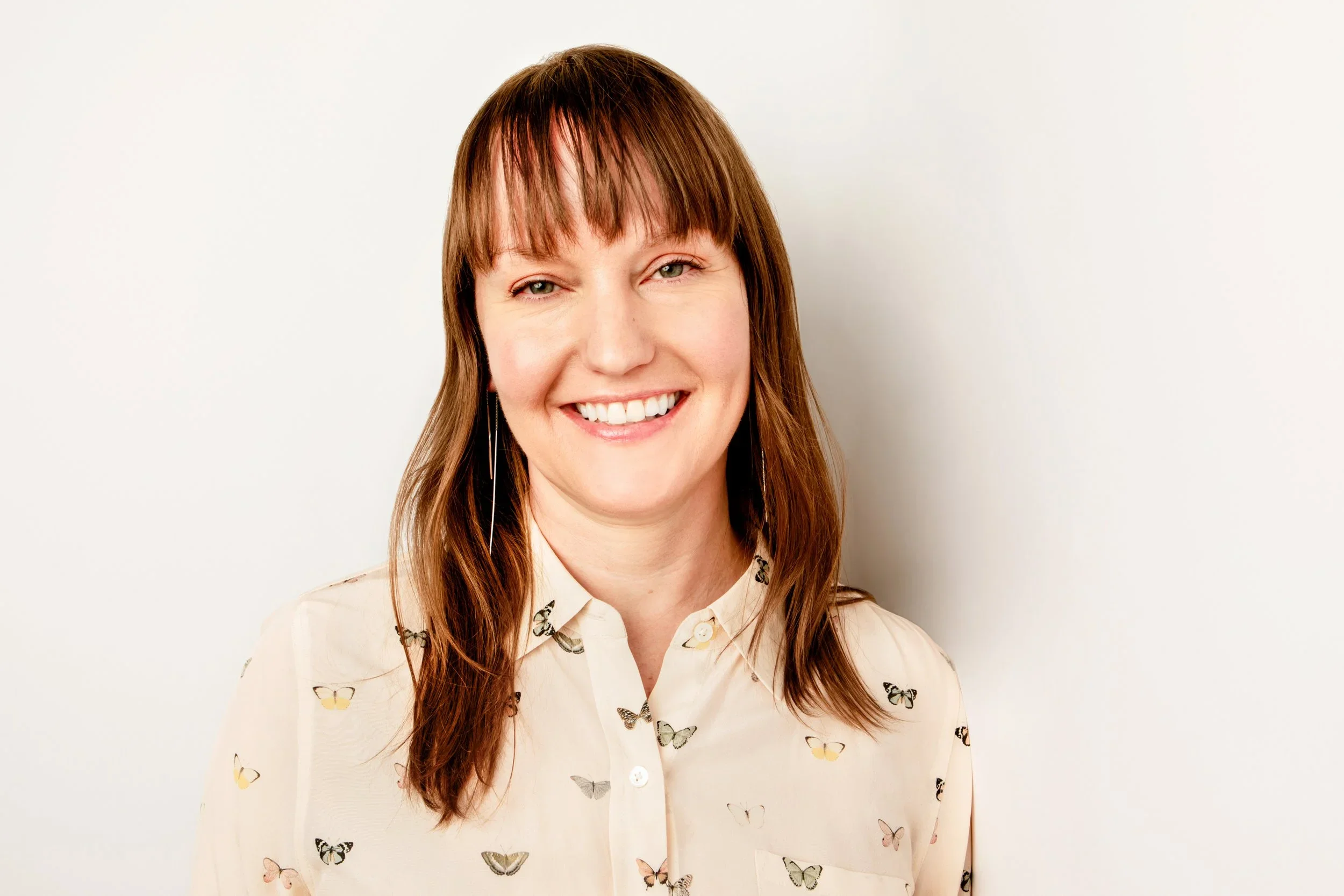 A woman with long brown hair, wearing a cream-colored blouse with butterfly print, smiling against a white background.