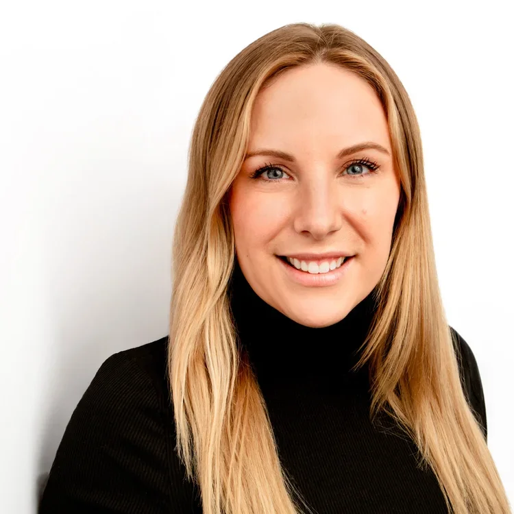 Close-up portrait of a young woman with long blonde hair and blue eyes, smiling against a white background, wearing a black top.