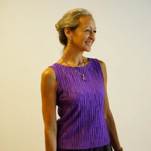 Smiling woman with gray hair styled back, wearing a sleeveless purple top, gold jewelry, and standing against a plain beige background.