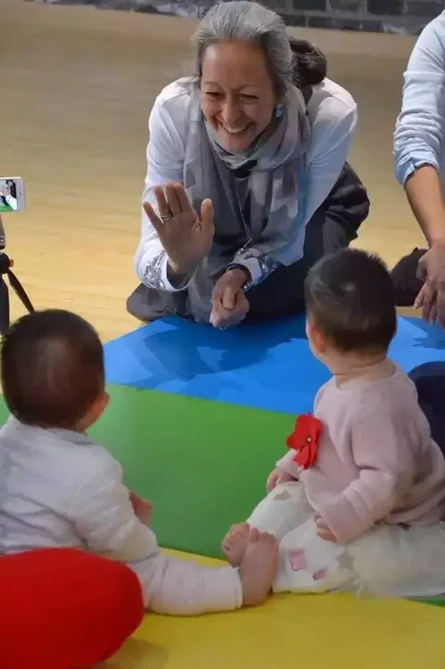 A smiling woman crouches on the floor waving at two babies sitting on a multicolored mat. The babies are facing away from the camera, engaging with the woman.