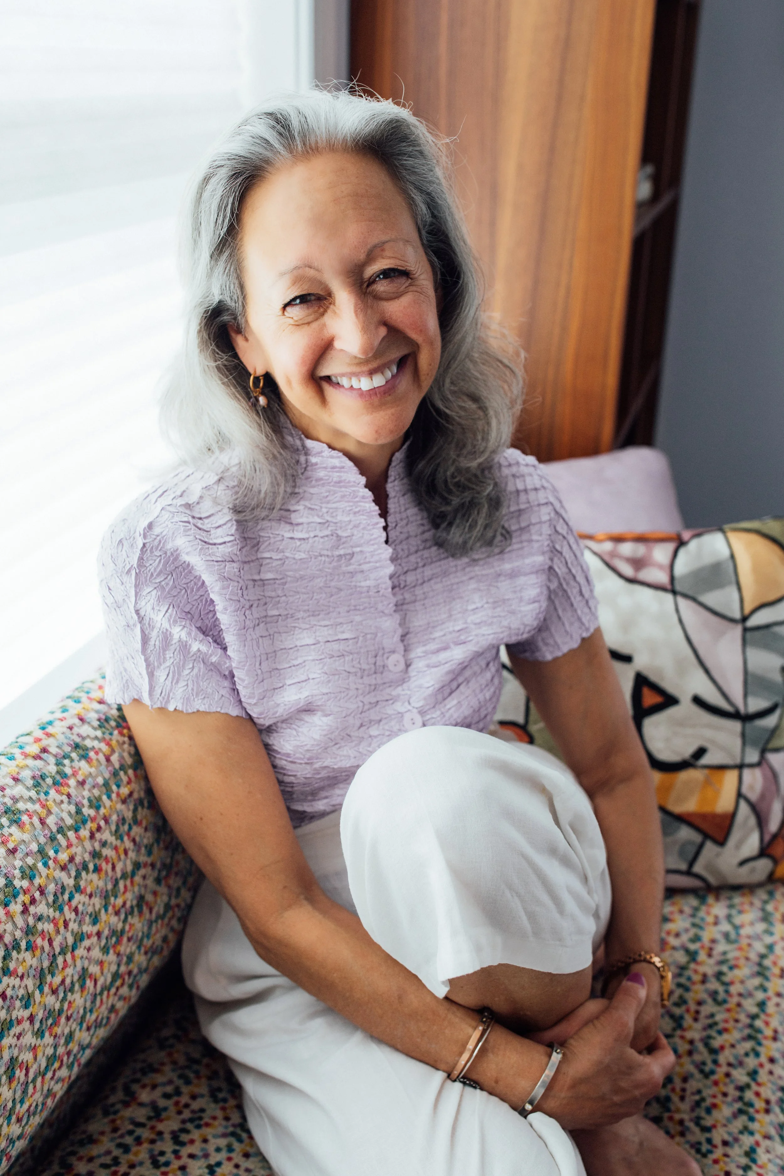 A smiling older woman with gray hair, wearing a lavender blouse, sitting on a colorful couch, holding a white pillow, in a well-lit room with a wooden wall behind her.