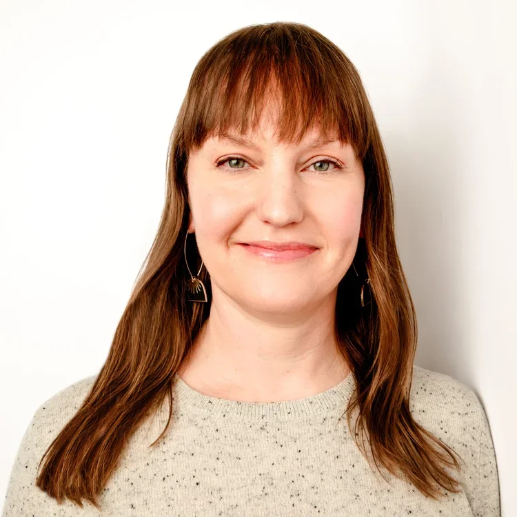 A woman with long brown hair and bangs, smiling, wearing a beige sweater with small black speckles and gold geometric earrings, against a white background.