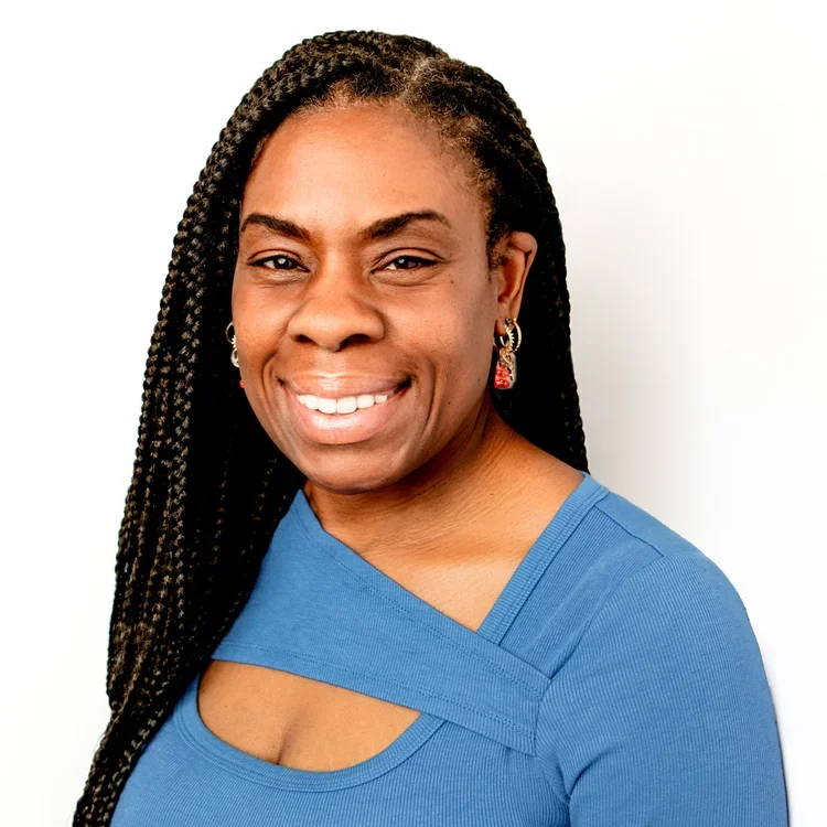 A woman with long braids smiling, wearing a blue top and earrings, standing against a white background.