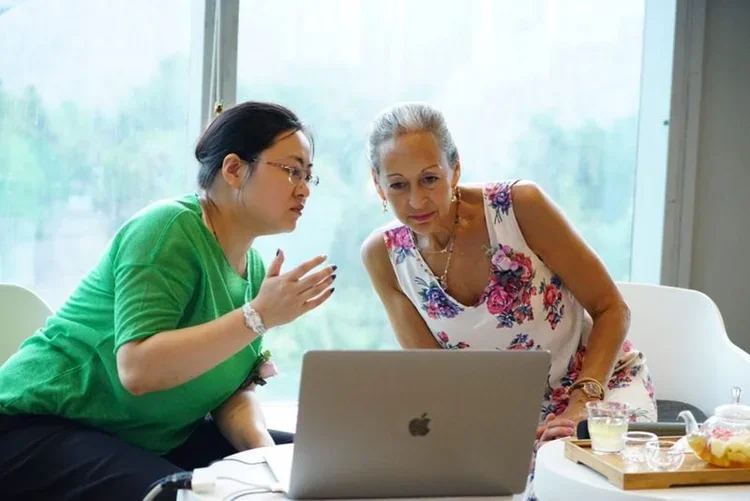Two women looking at a laptop, one in a green shirt and glasses, the other in a floral dress, sitting at a table with snacks and drinks in a bright room with large windows.