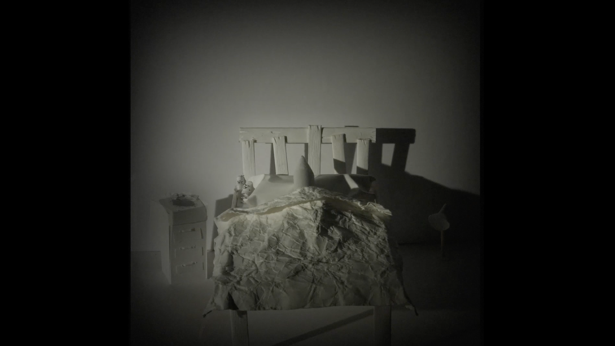 A dimly lit bedroom with a white wooden bed, a wrinkled bedspread, and a small white nightstand with items on top. The room appears to be mostly empty and shadowy.