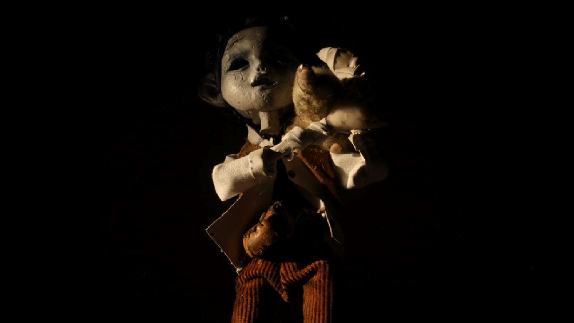 A doll with a cracked face, wearing a white shirt and brown pants, is lying on a dark background with a single light source highlighting its face and upper body.