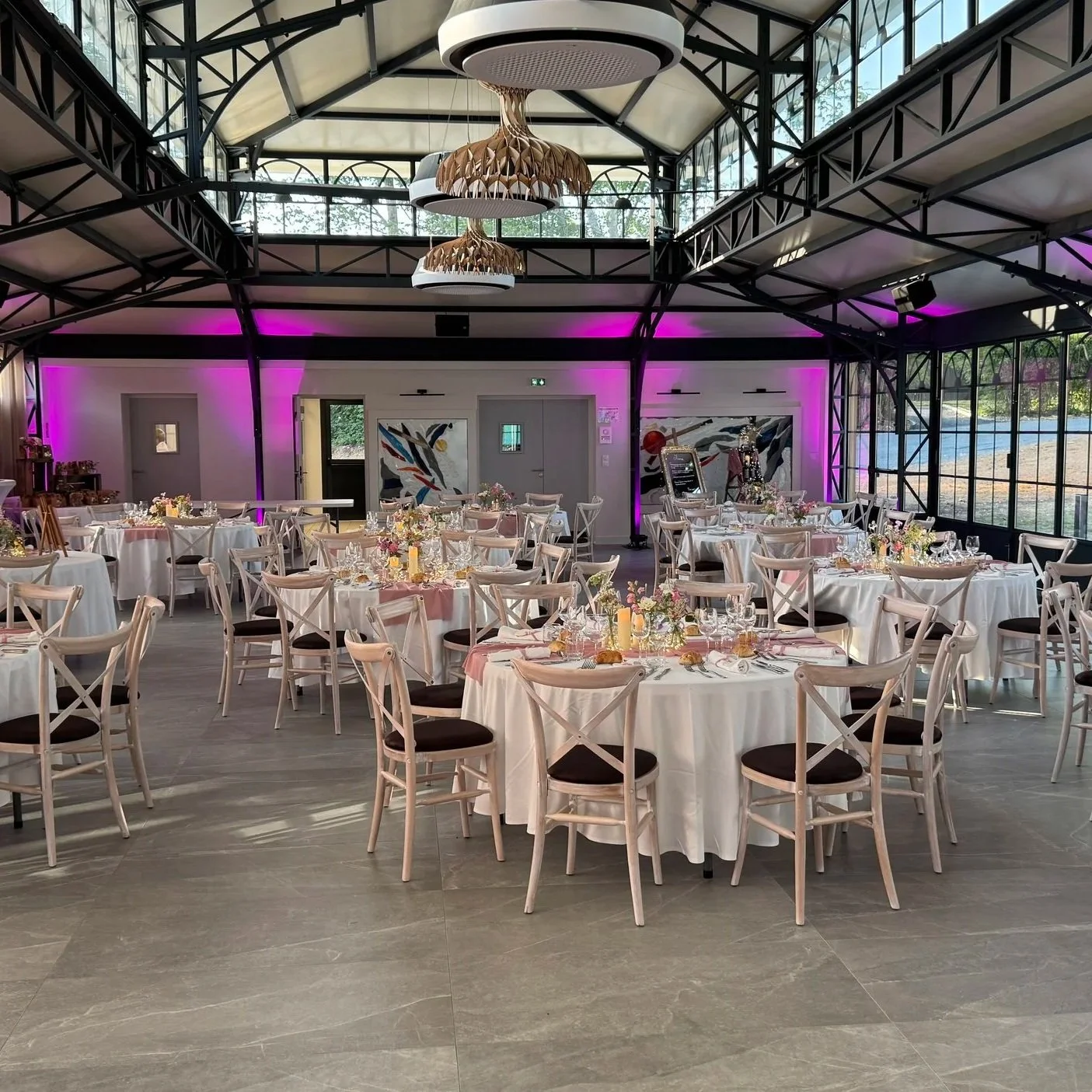 Salle de réception élégante avec des tables rondes habillées de nappes blanches, décorées de fleurs et de bougies, entourées de chaises en bois clair avec assises foncées. Des lumières violettes illuminent la pièce, qui a un plafond en structure métallique avec des luminaires modernes en bois.