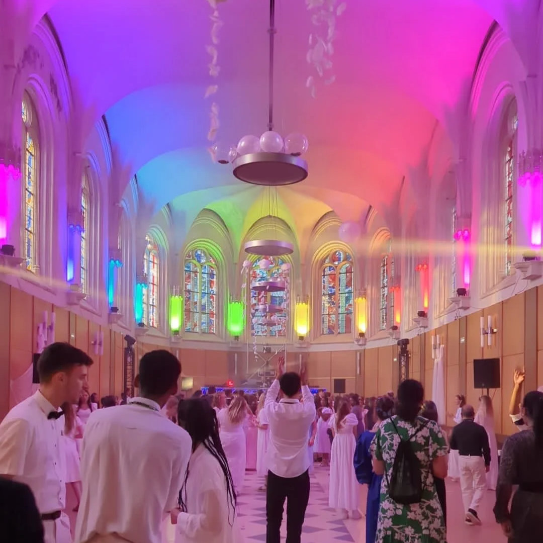 Une grande salle de bal avec un plafond en voûte, décorée de lumières colorées dans des tons de violet, bleu, vert, jaune, orange et rouge. De nombreuses personnes habillées en blanc ou en tenues de soirée participent à une fête ou un événement, avec des vitraux colorés en toile de fond.